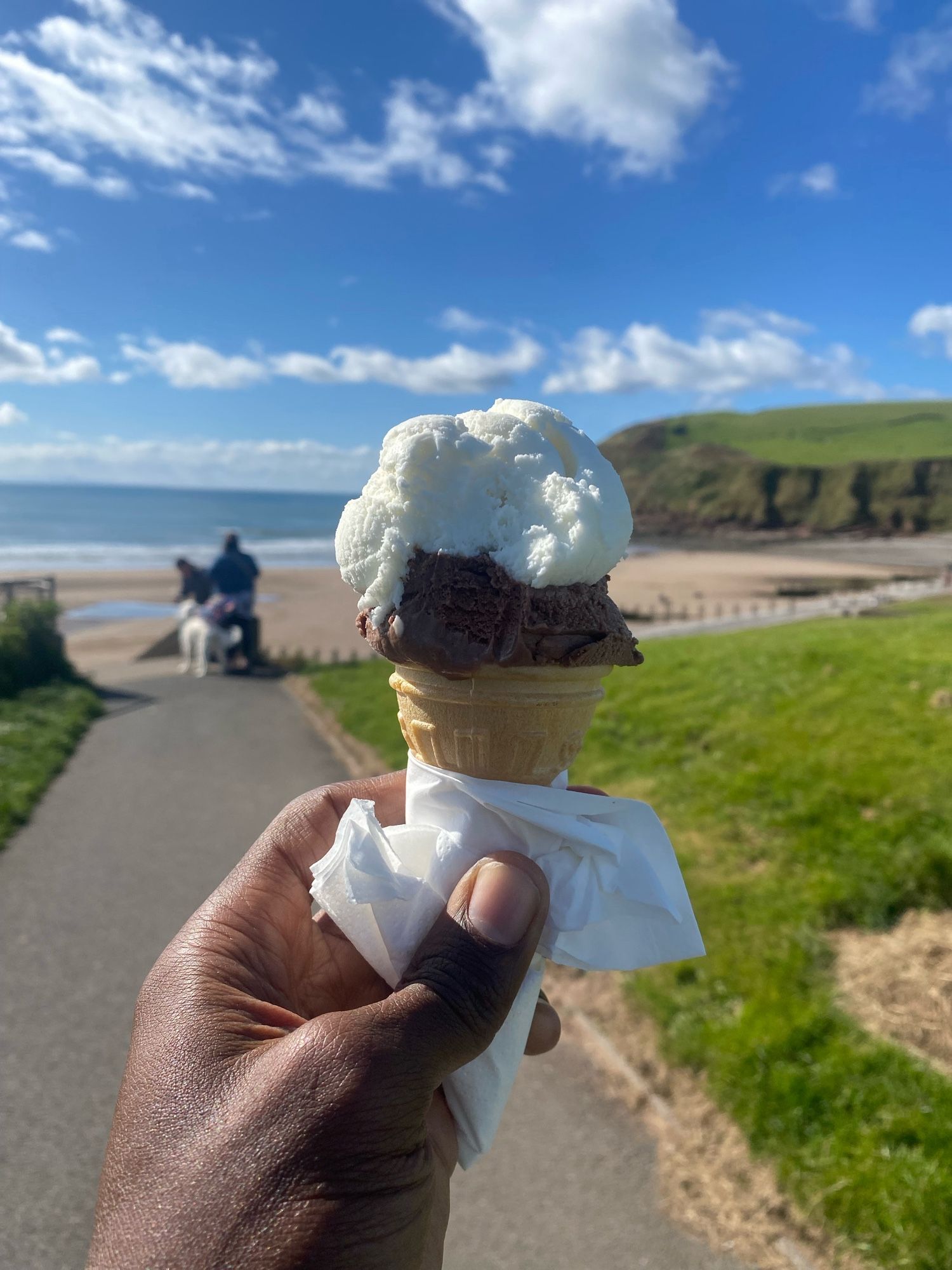 My outstretched hand holding an ice cream. Out of focus, in the background, is a similar beach scene to that in the first image.