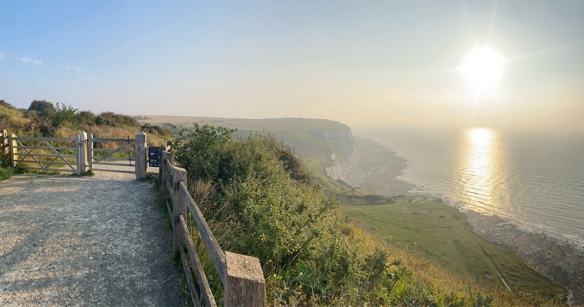 A cliff top view out to see below a rising sun. There is a gravel path in the foreground and a gate.