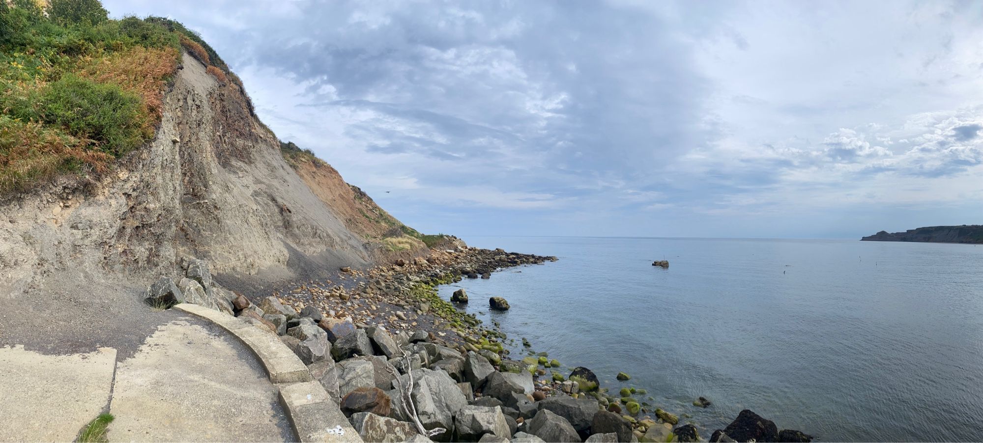 A view along a low, grey mudstone cliff line , capped by grass. The sea is calm and sits below a cloudy sky. The shoreline is rocky.