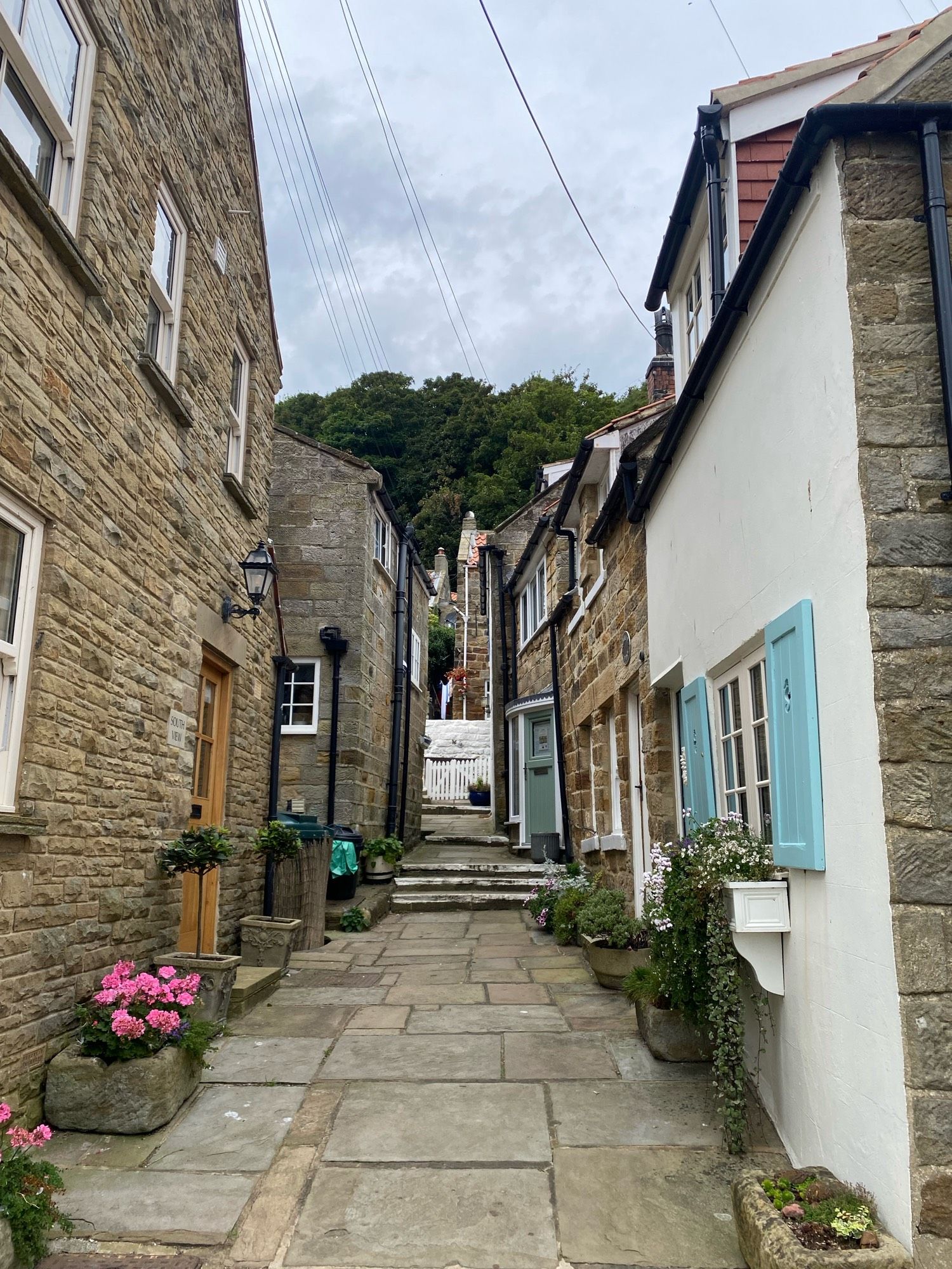 A view down a narrow passageway between light-brown very small, very pretty sandstone cottages. The sky is cloudy and there are trees behind the houses at the top of the passage.