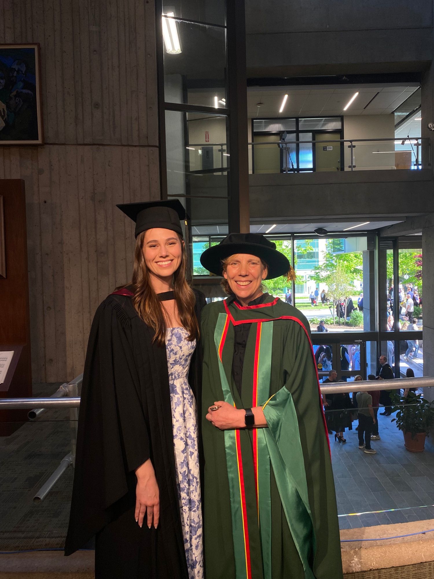 Two women wearing academic robes and hats are standing on a balcony inside a building. They are smiling at the camera. In the background are more people chatting and a window with a view outside onto trees, buildings and sunshine.