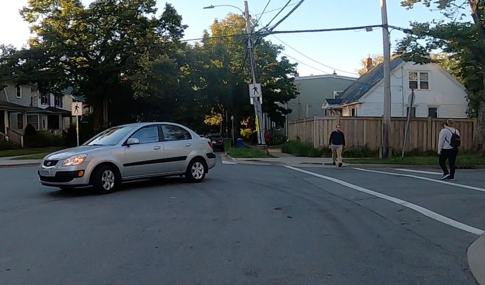 An image of a street from the POV of a cyclist. A silver grey car has just pulled out from a side street in front of the cyclist, moving from the right side of the image to the left and forcing the cyclist to brake. There are two people crossing the side street who were also trying to safely. There are trees and houses in the background of the image and the early morning sky is a cloudy blue with signs of the rising sun.