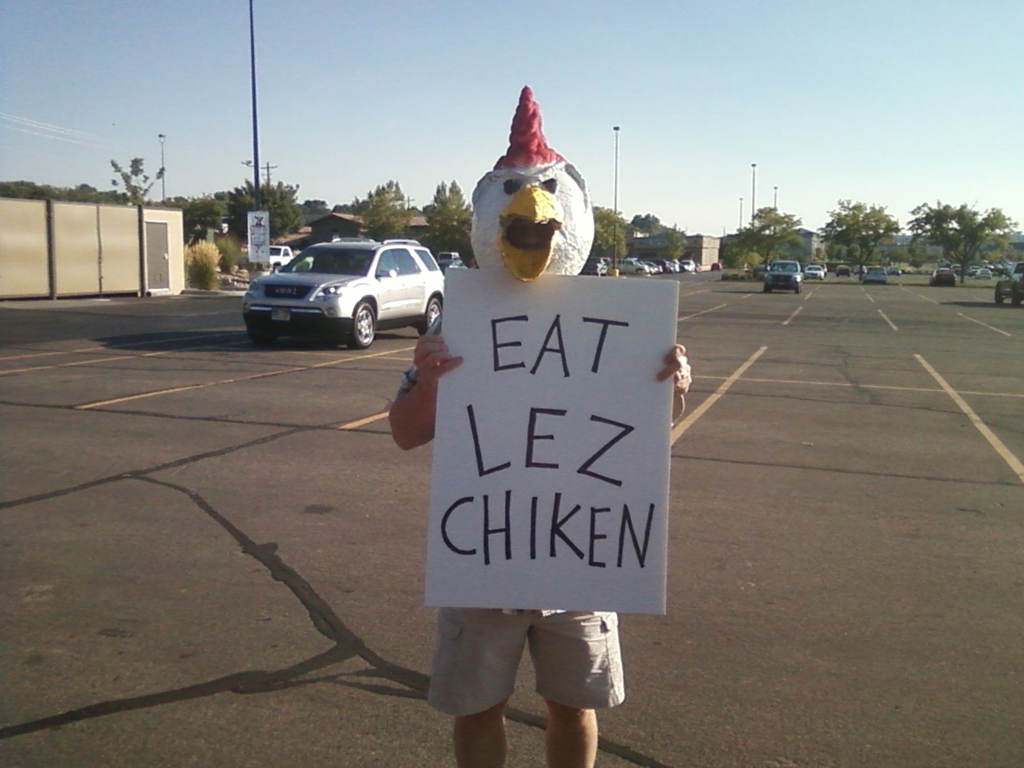 A man wearing a papier-mâché chicken head and holding a sign that says "eat lez chiken" in a parking lot.