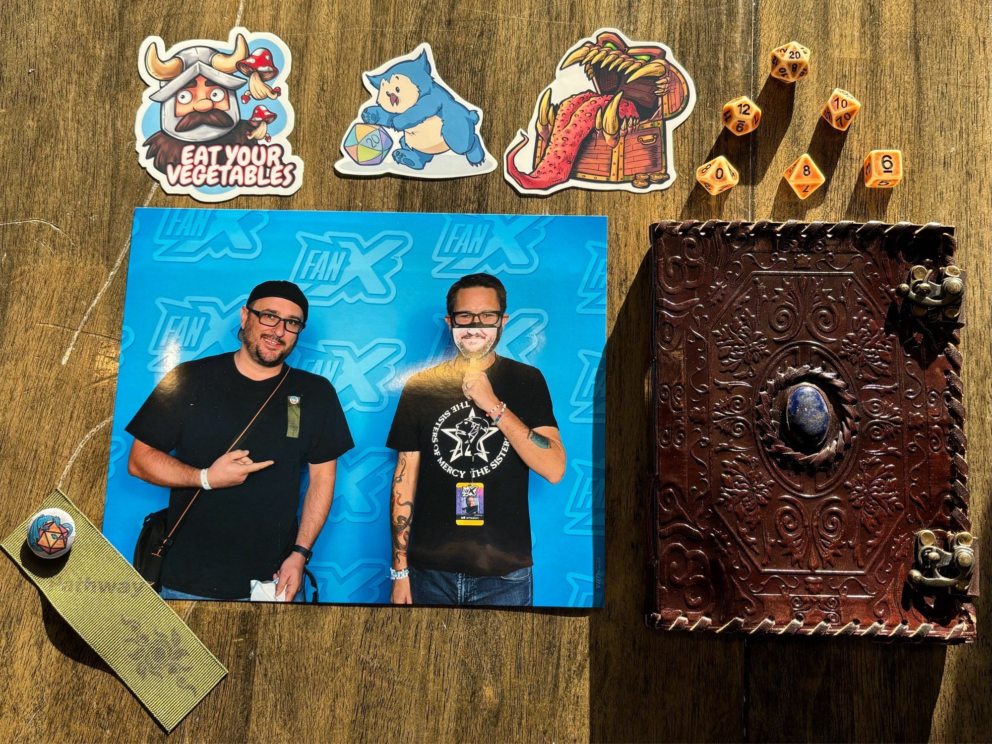 Three stickers, left to right: Senshi, the dwarf warrior from Delicious In Dungeon; an owlbear chasing a 20-sided die; and an angry treasure chest mimic with its tongue sticking out. A set of tan, weathered polyhedral dice. A photo of me and Wil Wheaton along with my "Nature's Pathway" ribbon and We Geek Together pin from the convention game, and a pleather notebook with latches and a gem at the center of the intricate designs on the cover, made to appear old and mystical.