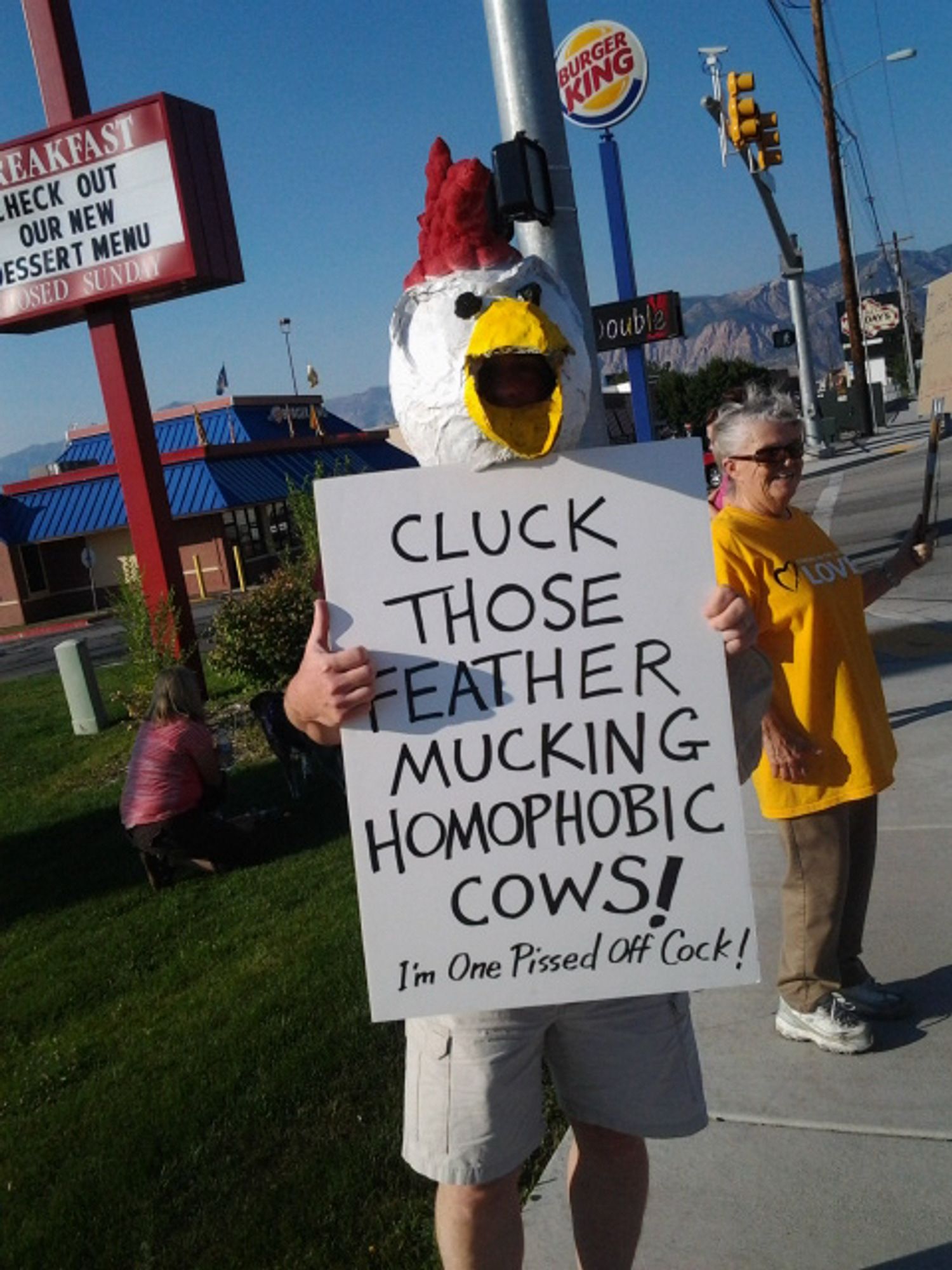 A man wearing a papier-mâché chicken head and holding a sign that reads "CLUCK THOSE FEATHER MUCKING НОМОРНОВІС COWS! I'm One Pissed Of Cock!" He is standing on a sidewalk in front of a Chik-fil-A restaurant. Photo from August 2012.