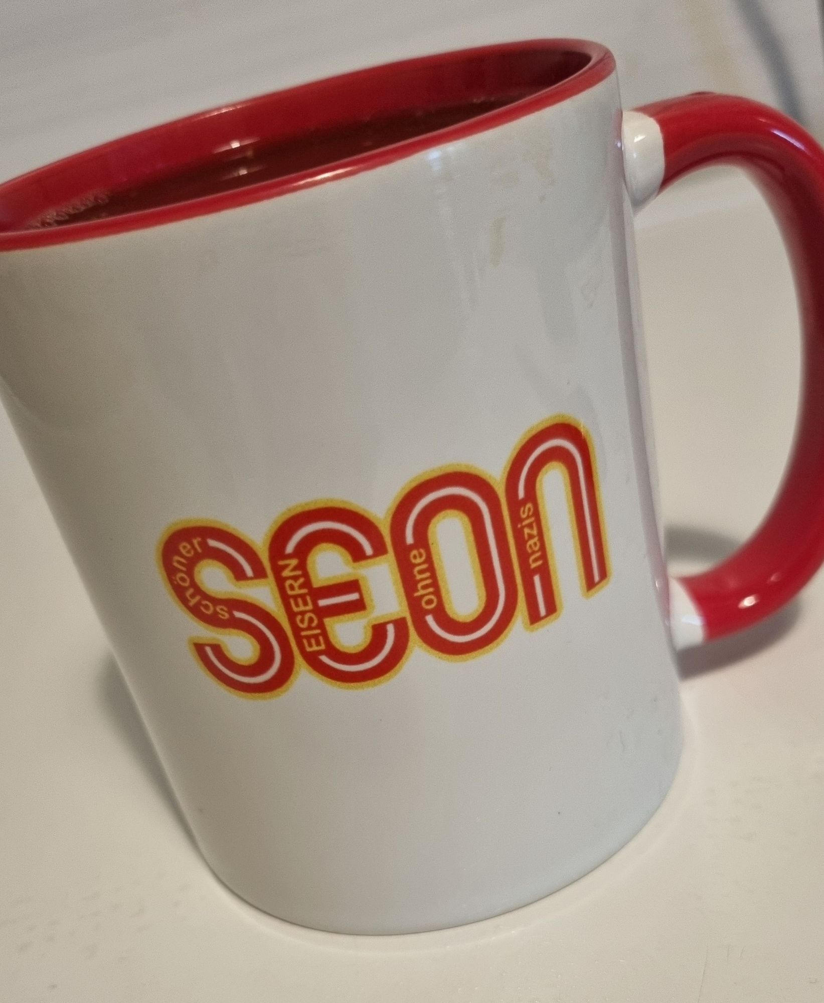 Weiße Tasse mit rotem Henkel und roter Innenseite. Aufschrift der Tasse "SEON" (Schöner Eisern ohne Nazis).