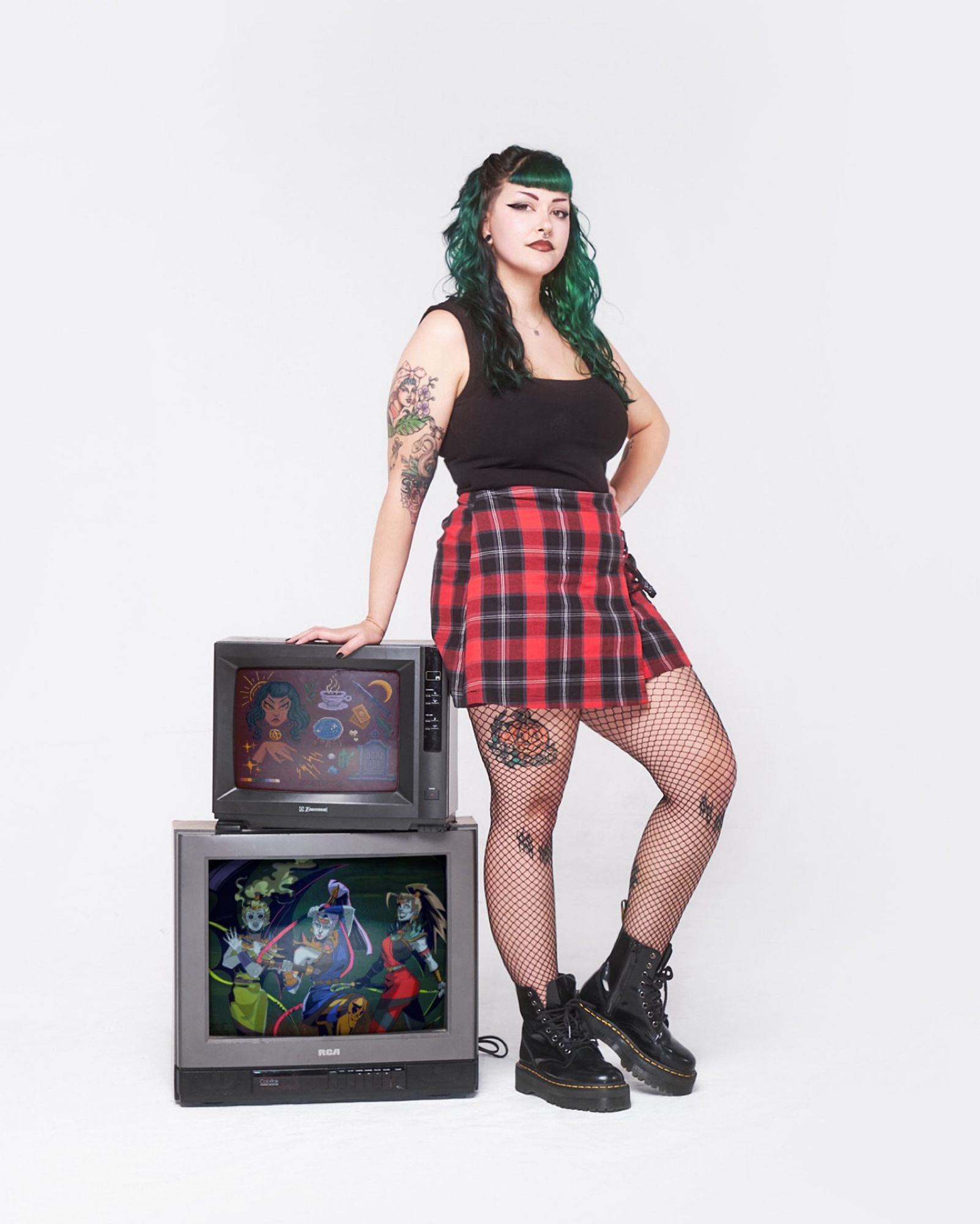 Photo of Zoe lTreez standing in a white background beside two CR TVs. Displayed on the TVs are two different art works done by ZoeTreez. The tope TV displaying a pixel art drawing and the bottom being a Fury Sister fan art. Treez is in a black top, plaid skirt, fishnets and doc boots. Treez hand is rested on the tvs and the other in her hip. Treez has long green hair and sharp eyeliner.