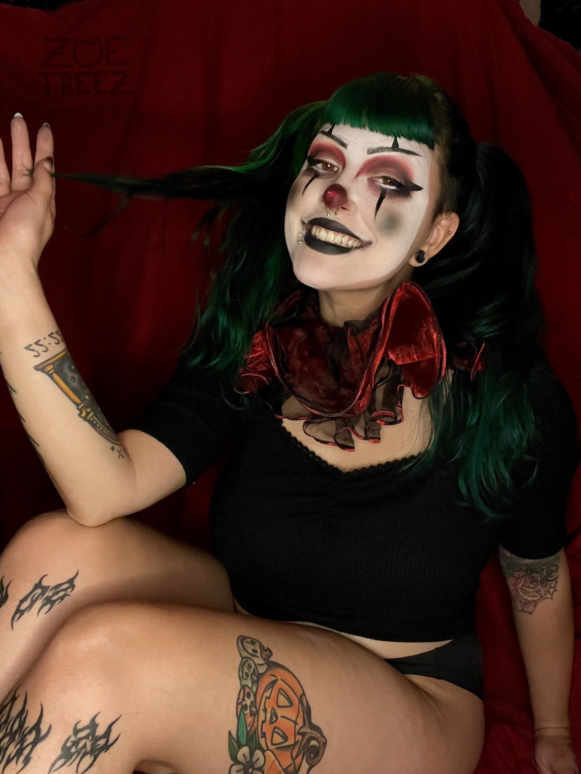 Treez (green haired fem) with black, white and red clown make up. They have a black and red ruffled collar and are sitting in front of a red back drop. She’s smiling like she’s going to bite your ankles in your sleep