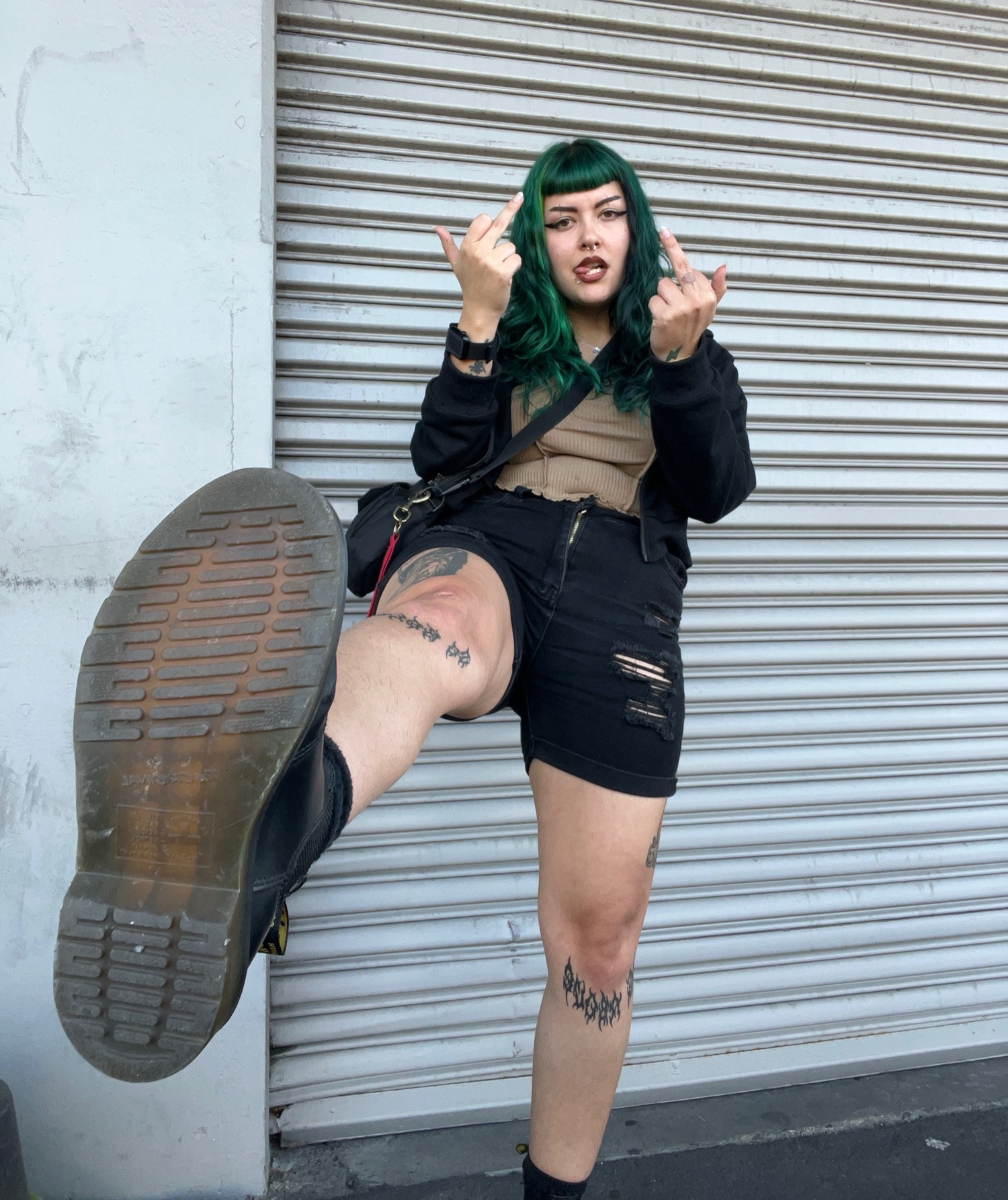 Picture of Treez standing in front of a grey wall. Treez is posed with one leg up, kicking their boot up to the camera. You can see the underneath of her docs as if she’s about to kick you in the face. She’s wearing distressed black shorts, a brown tank and black zip hoodie. Her middle fingers are up and her tongue is sticking out to the side of her mouth. She’s got black winged eyeliner, sick tattoos and epic green hair