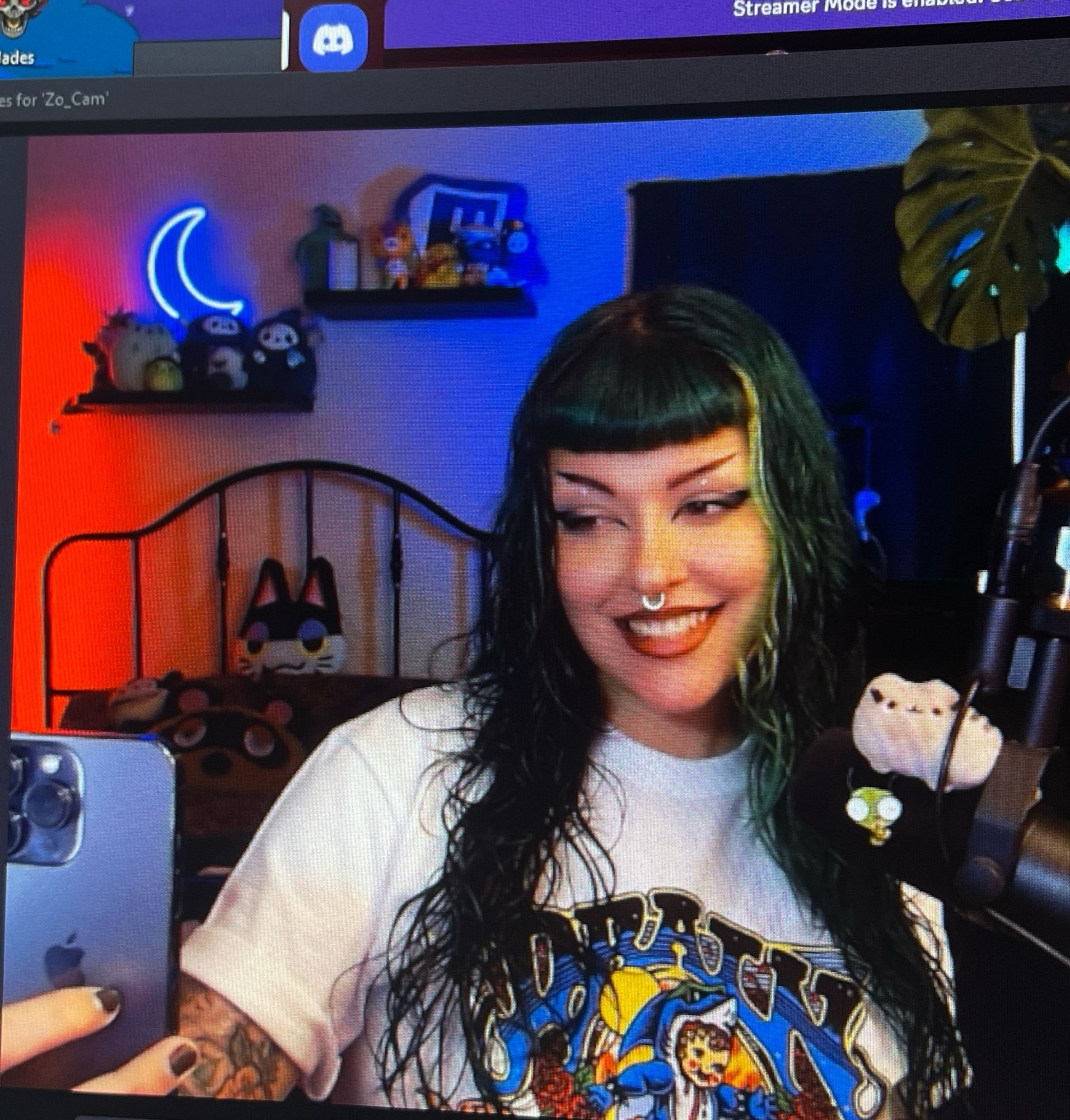 A photo of Treez, a femme with long green hair and bangs. They have a septum ring and a DRAIN band tee on. The photo is of the webcam view on their computer. You can see neon lights and plushes in the background and their cute lil pusheen and gir on the microphone