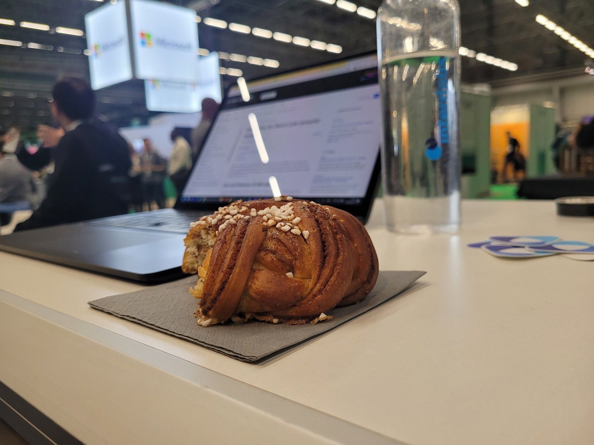 A picture of a desk with a cinnamon bun in front and a laptop and people blurry in the background