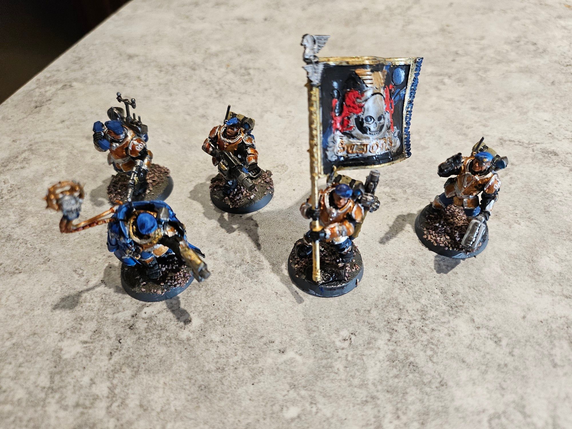 A squad of 25mm figurines from Warhammer 40,000. These are painted in bronze with silver highlights.  One trooper has a banner in gold black and red. The commander has a long blue overcoat.