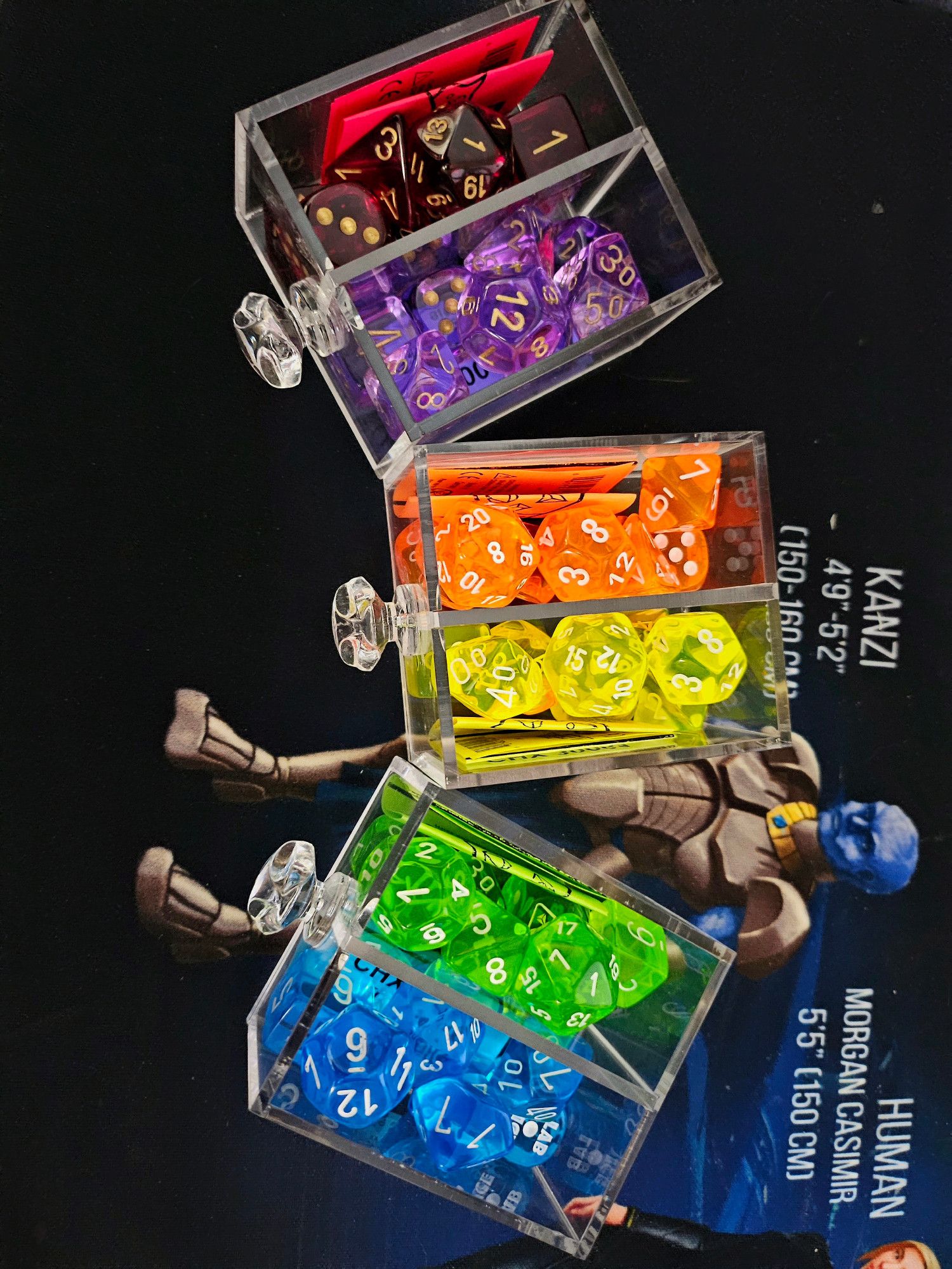 Six sets of dice in three drawers. All are Chessex sets from the 2023 wave of lab dice.
Top is transparent crimson dice with gold numbers.
Next is transparent lavender with gold numbers.
Then transparent neon orange with white numbers, then transparent neon yellow with white numbers.
Bottom drawer is a very neon transparent green (called 'Rad Green' by Chessex) with white numbers and a transparent blue with white numbers.
Background is one of the aliens from the Duchy of Terra series on a mousepad that isn't for sale.