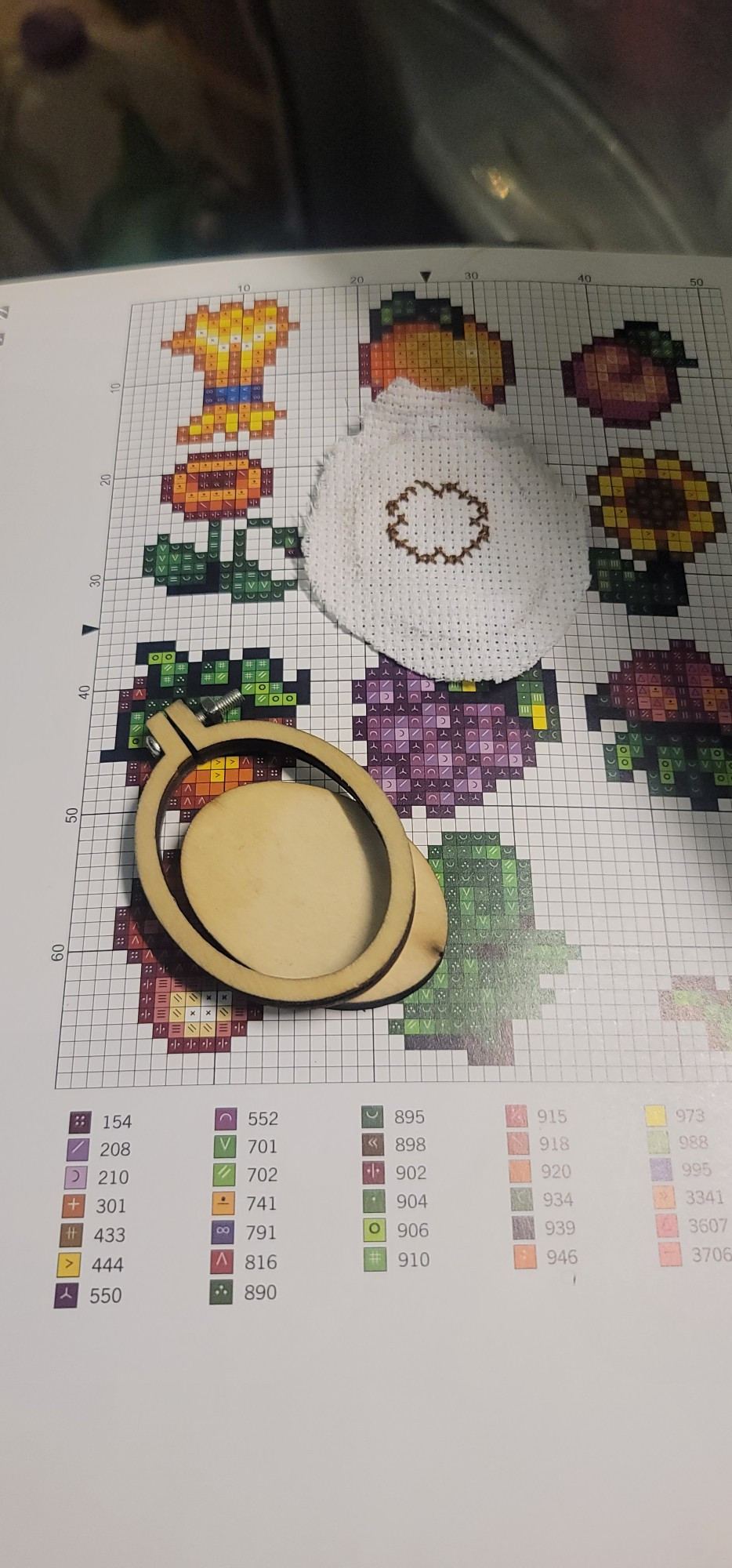 A photo of a cross-stitch pattern with various Summer crops in Stardew Valley, with a wooden keychain hoop and an incomplete stitched sunflower.