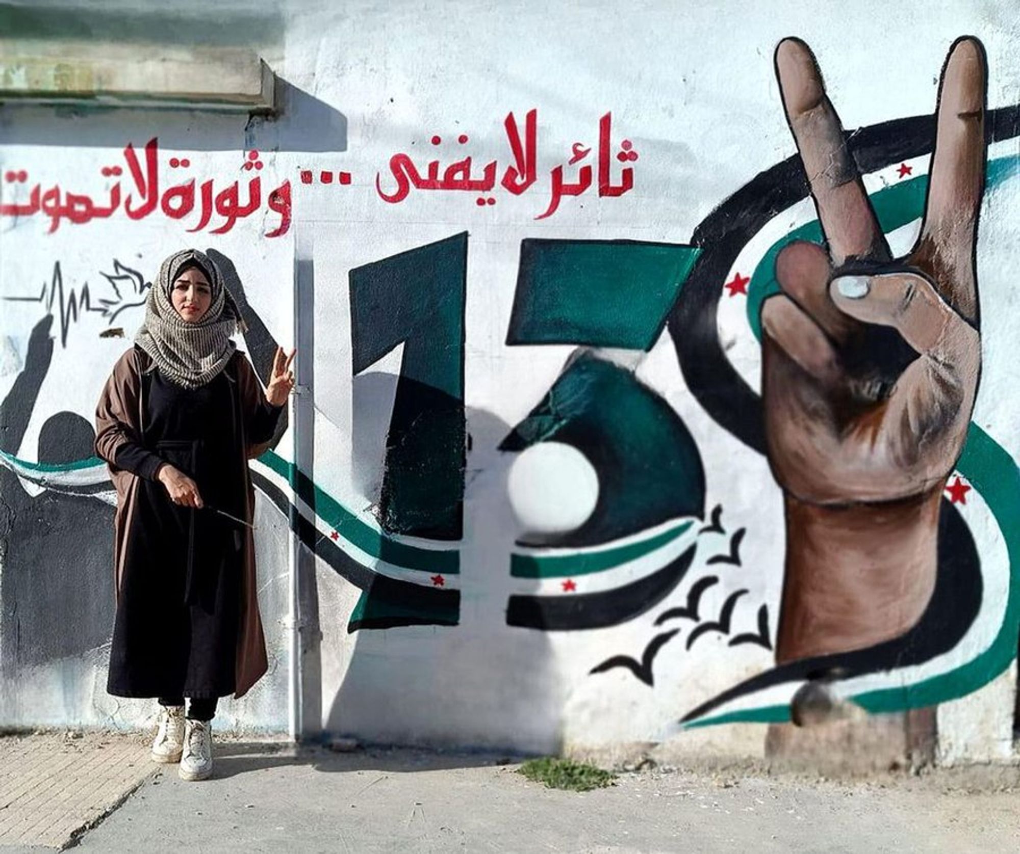 Picture of Salam standing infront of her mural: 13 years is painted in the colours of the revolution flag, as well as a 'V' for Victory sign