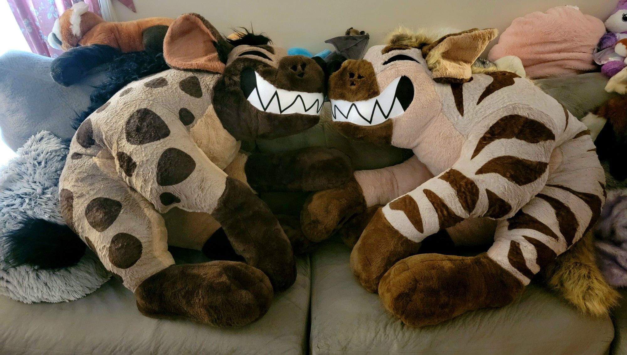 Two giant hyena plushies! Both are sitting on a couch, noses touching, mirroring one another. The hyena on the left is spotted, brown with dark brown spots, snout and feet. The hyena on the right is striped, tan with warm brown stipes, snout and feet. Both have their eyes closed and very large toony, toothy smiles.