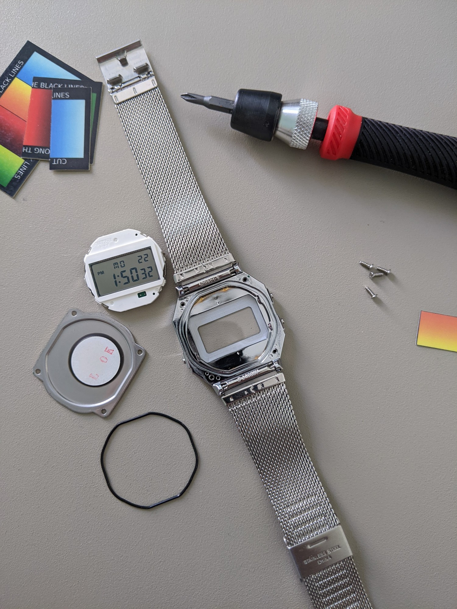 a disassembled silver casio digital watch, screws & digital display to the side along with a screwdriver and colored film