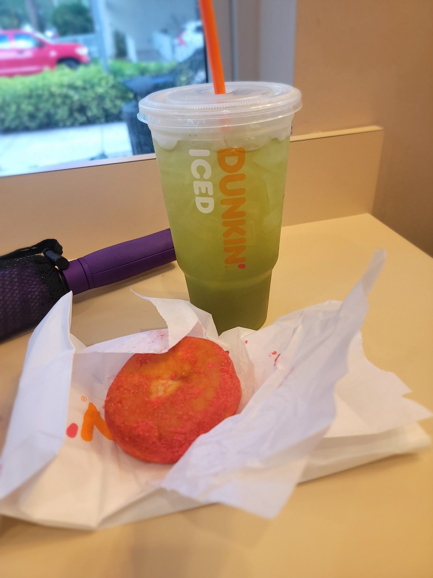 Taken inside a Dunkin donuts. 
On the beige table: 
To the left, part of a purple umbrella (the handle). 
In the center, a watermelon burst donut. A donut filled with watermelon jelly and dusted with watermelon flavored powdered sugar on the top. 
Behind the donut, a large Dunkin donut kiwi watermelon lemonade refresher. The drink is green in color with ice in it.