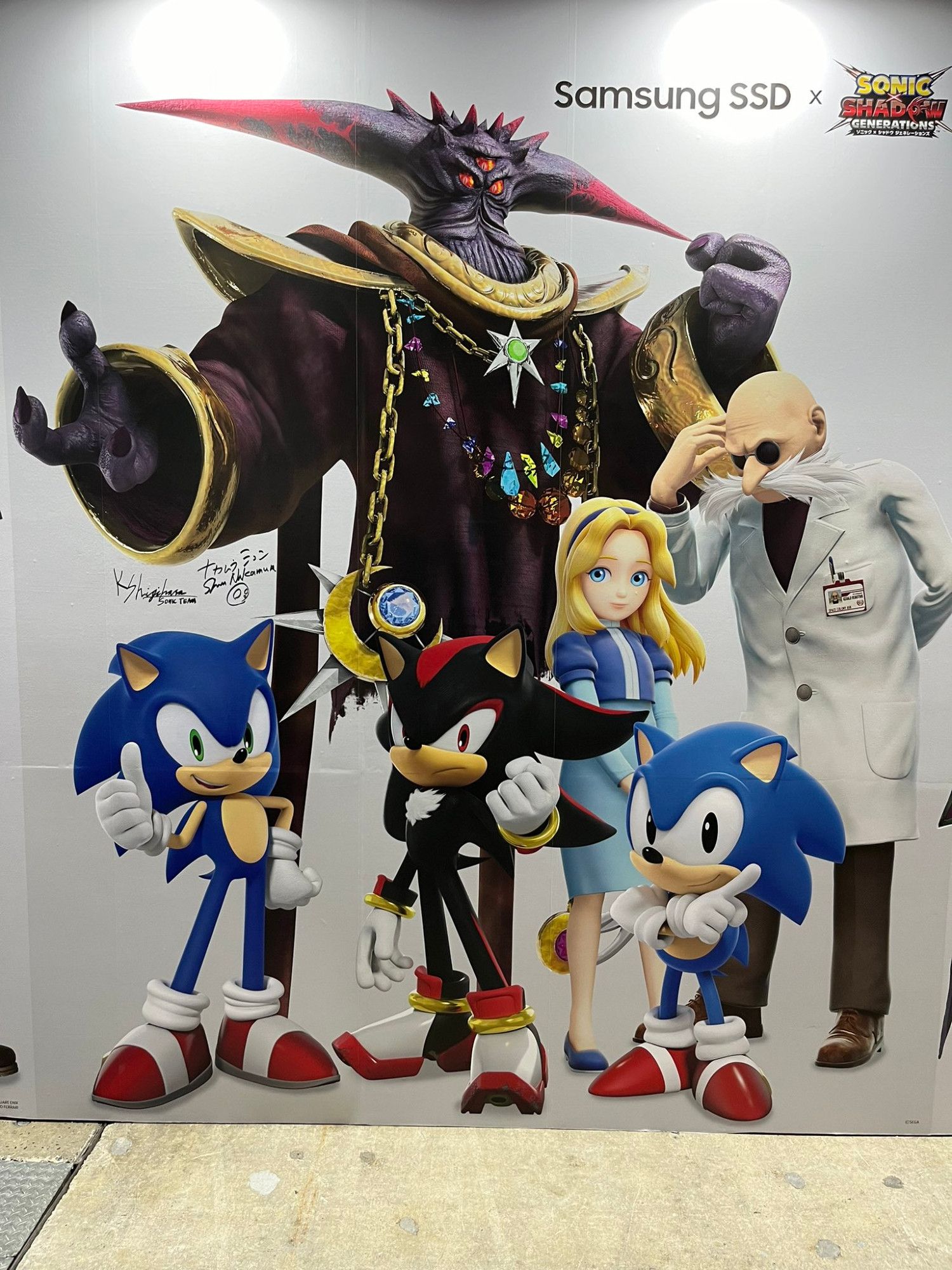 Photograph of a wall featuring several 3D renders made for Sonic x Shadow Generations but clearly not meant to all go together. There's Sonic smiling and giving an inappropriately-cheery thumbs-up, Shadow clutching his fist slightly menacingly, Maria with her hands together as her innocent hair is blown softly by passing space breezes, Black Doom looming ominously, and Professor Gerald Robotnik scratching his head in confusion as he happens to look like he's gazing down at Classic Sonic doing a pose like he's in a SEGA Genesis video game.