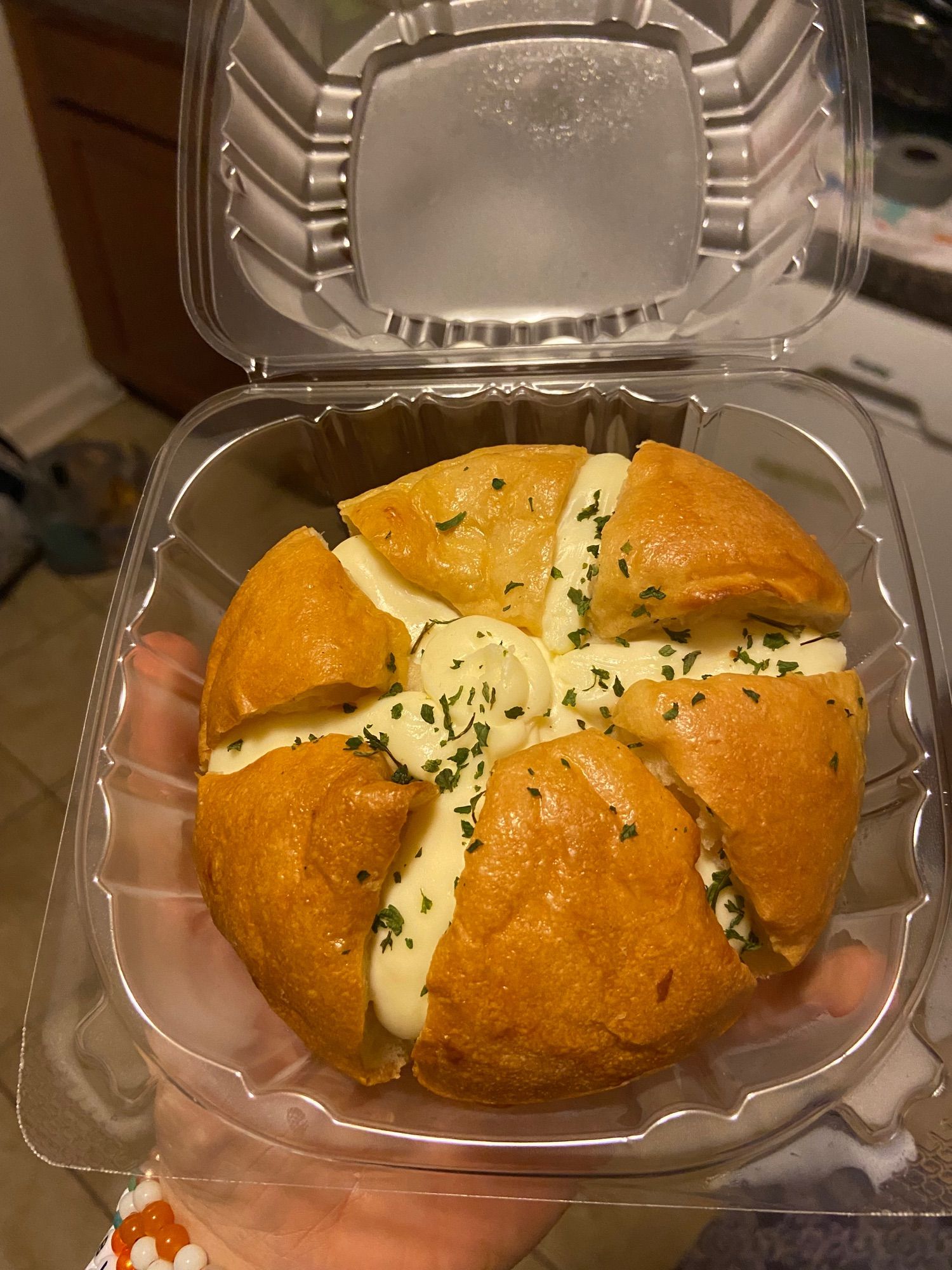 a clear plastic clamshell container holding a round, light-brown bread bun, which has been scored into six segments showing the filling within. the filling is a pale yellow cream cheese base flavored with honey and garlic, and sprinkled with some sort of finely minced green herb