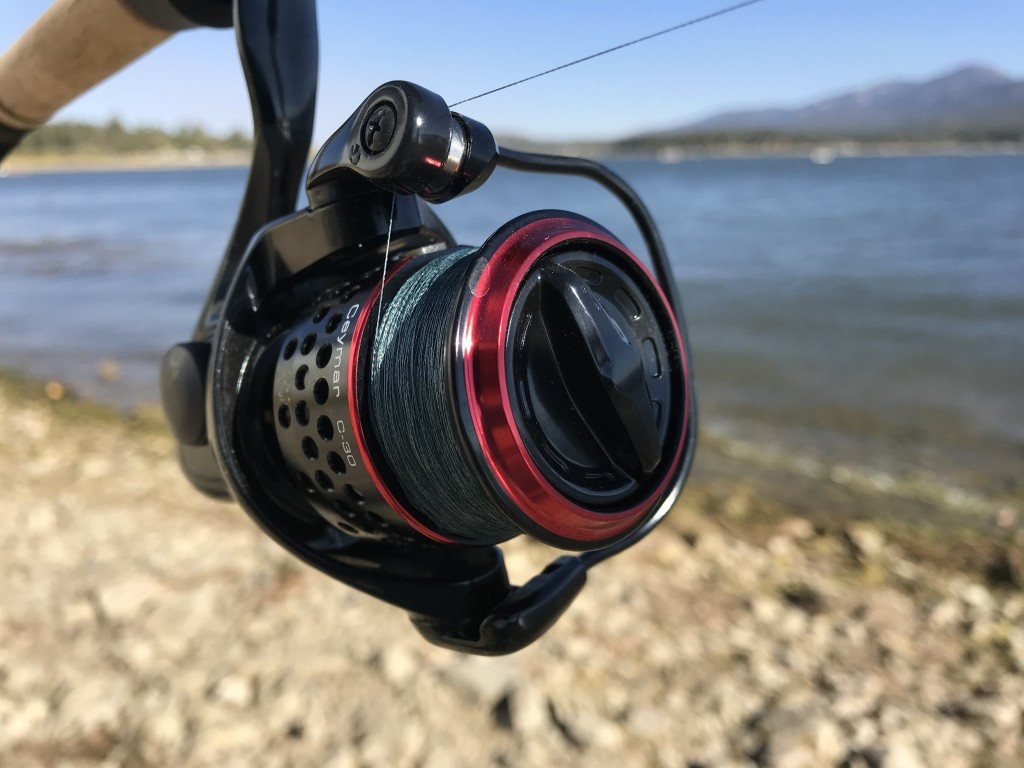 A photo of a fishing black and red fishing reel, taken against an out of focus stony beach with water and mountains in the background