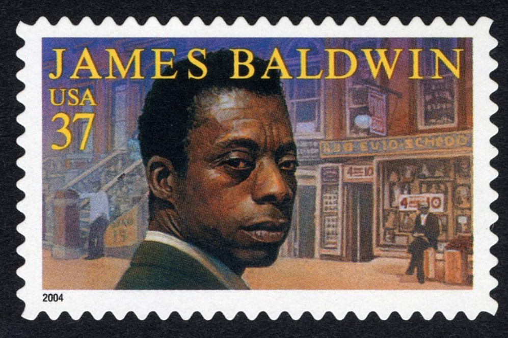 The James Baldwin stamp. Baldwin is depicted from the neck up looking over his shoulder in 3/4 profile. In the background is a brick city street with two Black men, meant to evoke Baldwin’s novel “Go Tell It on the Mountain.” 