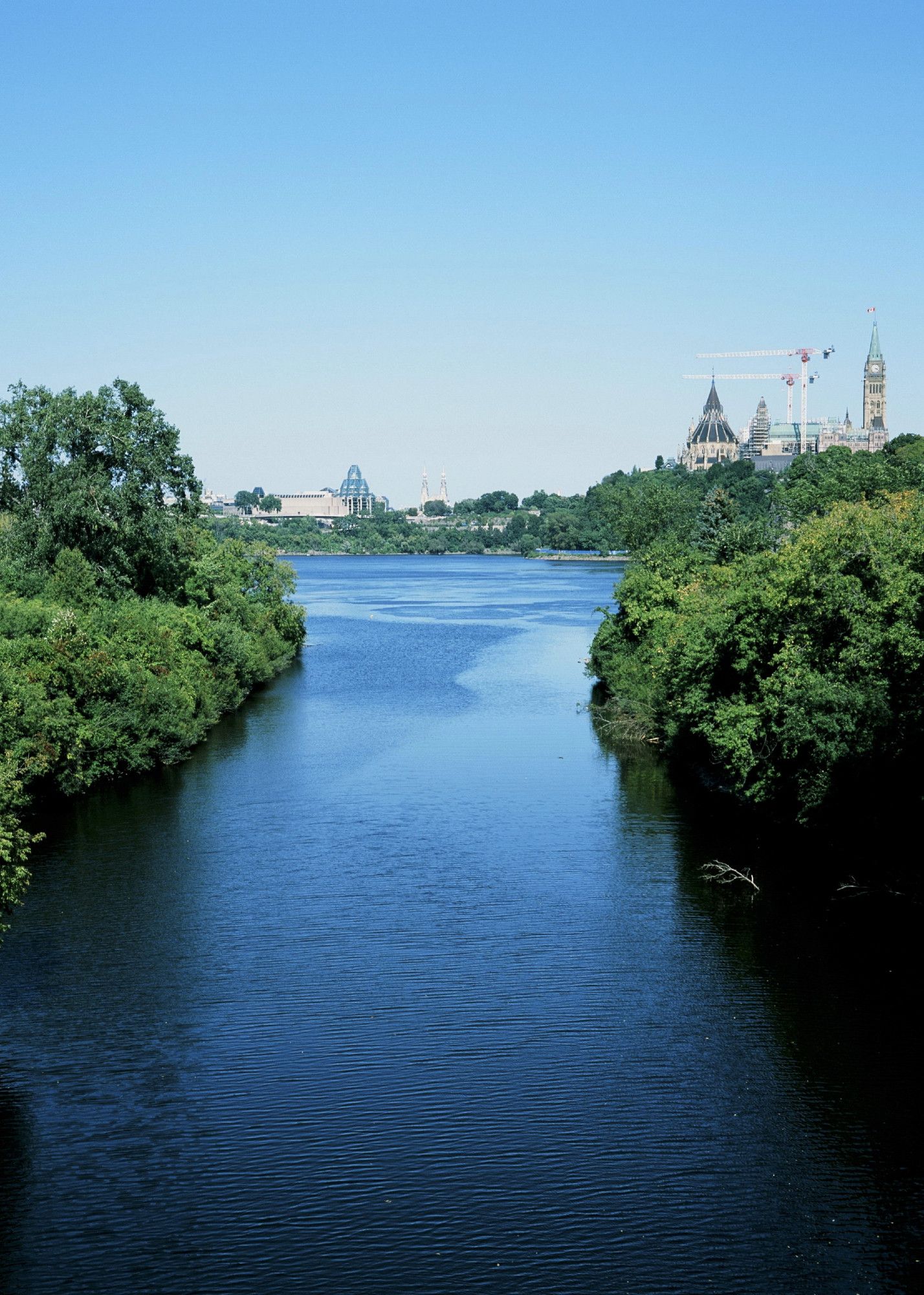 View of a small part of a larger river from a bridge. Trees and shrubs line the banks. In the distance, the neo-gothic Parliament buildings are visible perched on a rocky outcropping.