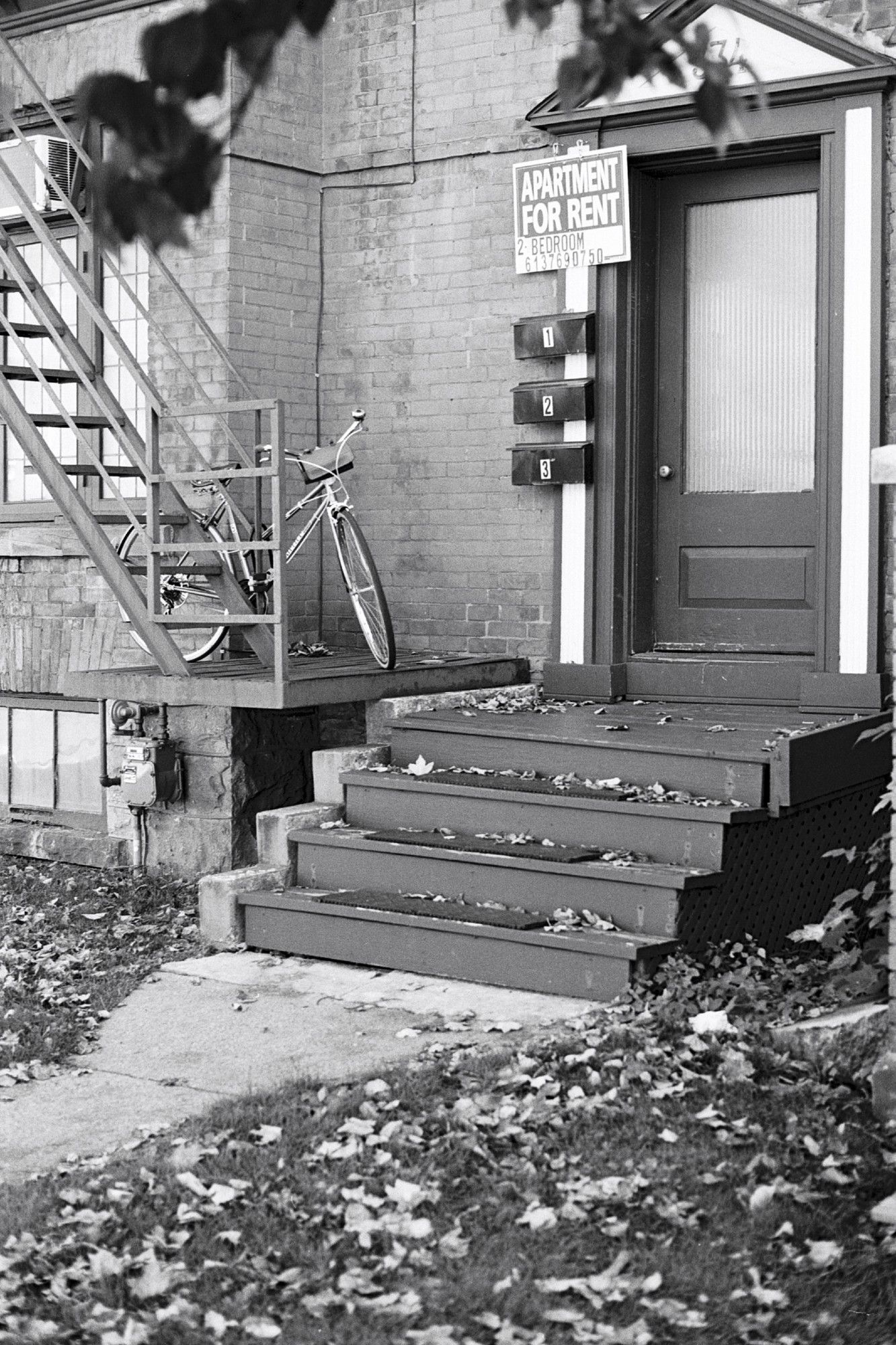 View of the entrance to upper-level apartments in a converted home. A sign affixed to the door frame advertises the availability of a two-bedroom apartment. A bicycle is locked to a fire escape.