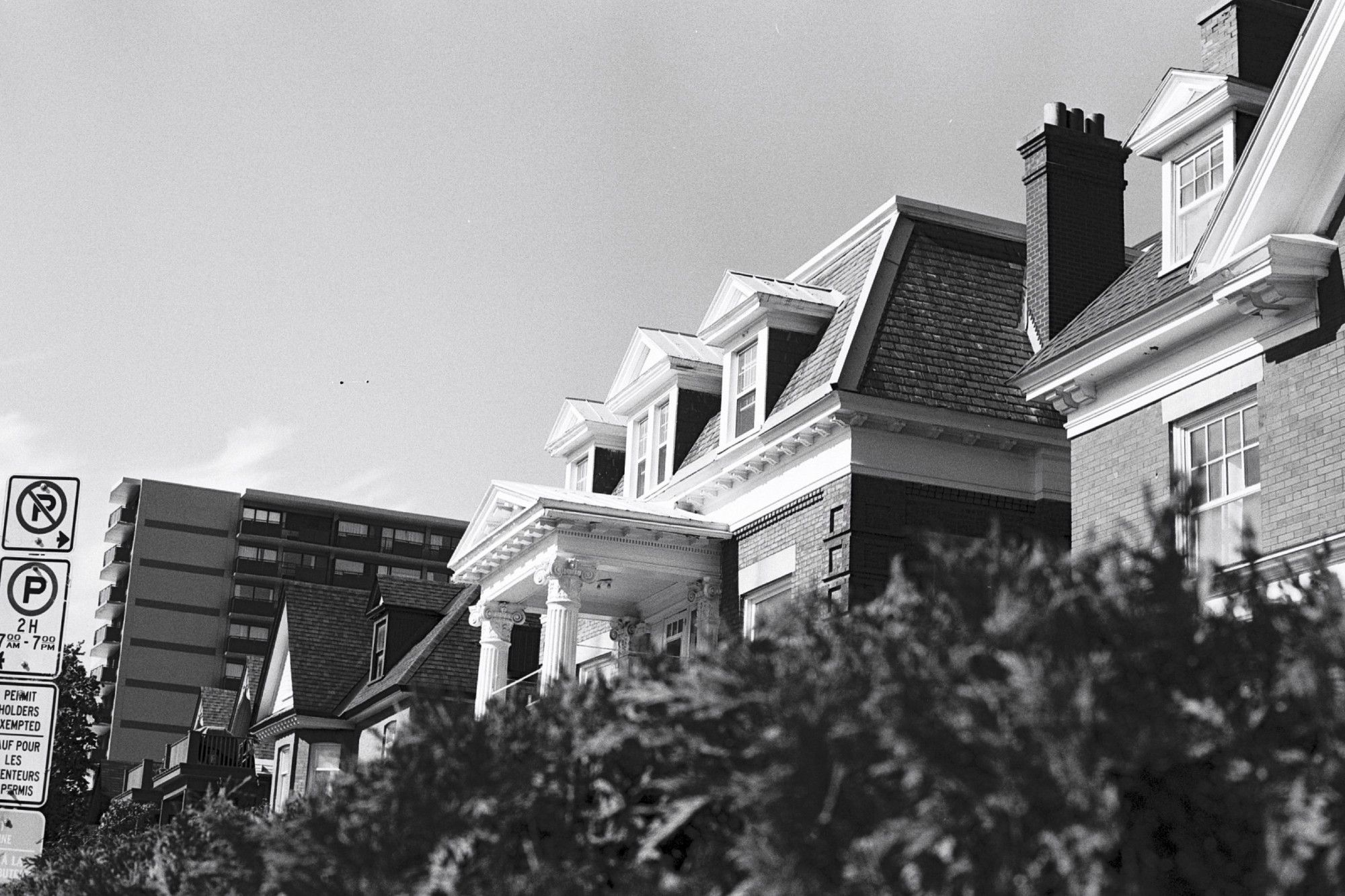 Low-angled view of a two-and-a-half storey home (converted into apartments). A tall cedar hedge in the front of its neighbouring property (to the right of the frame) obscures the home's first floor. The skies are bright and clear.