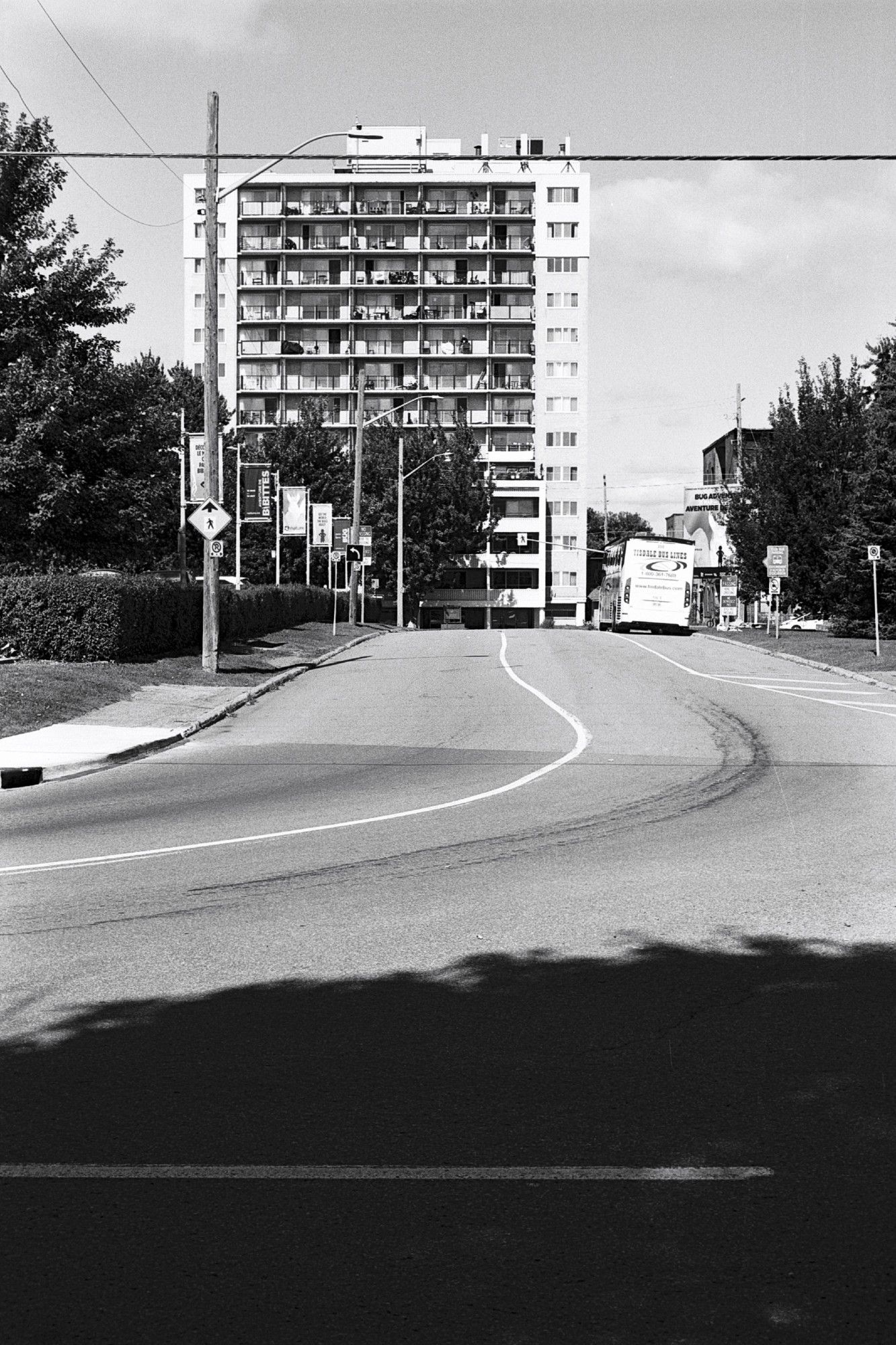 View down a road looking toward a midcentury apartment building. Tire tracks are visible on the curve.