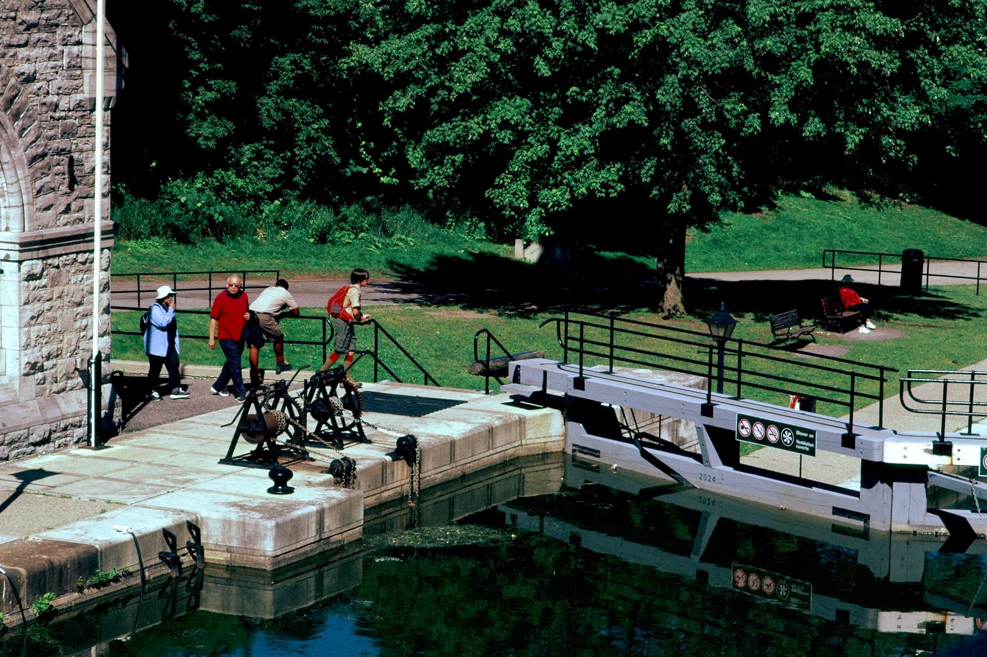 Tourists walking from behind a stone building to see one of the canal's grey-painted locks. An official with the locks' operations team leans on one of the railings watching the day.