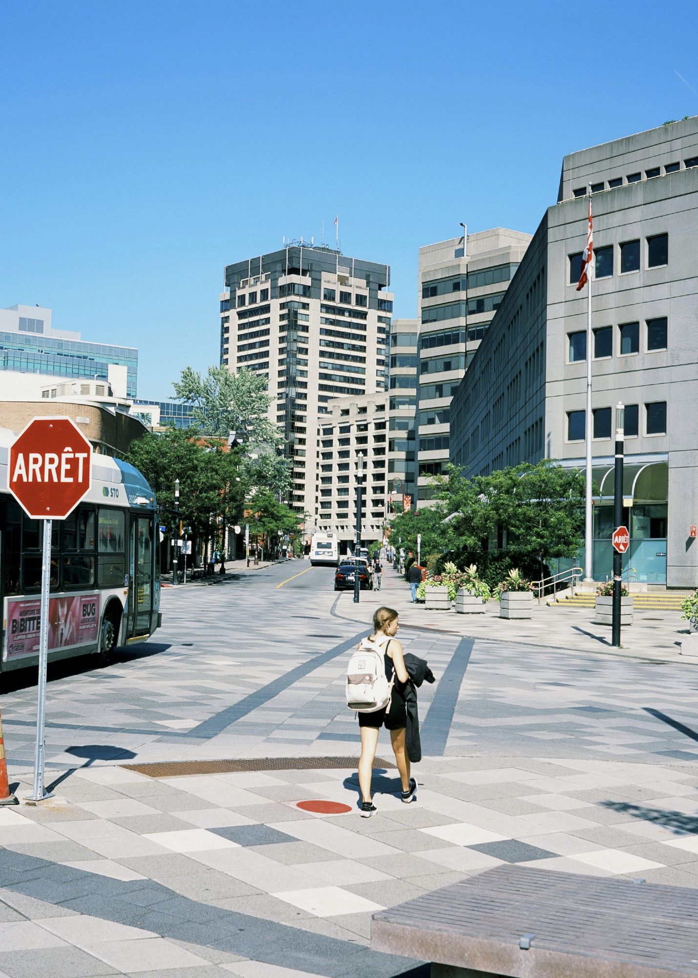 A woman wearing a backpack waiting to cross at an intersection. A city bus is passing to her left. A thicket of government office buildings line the right and into the background.