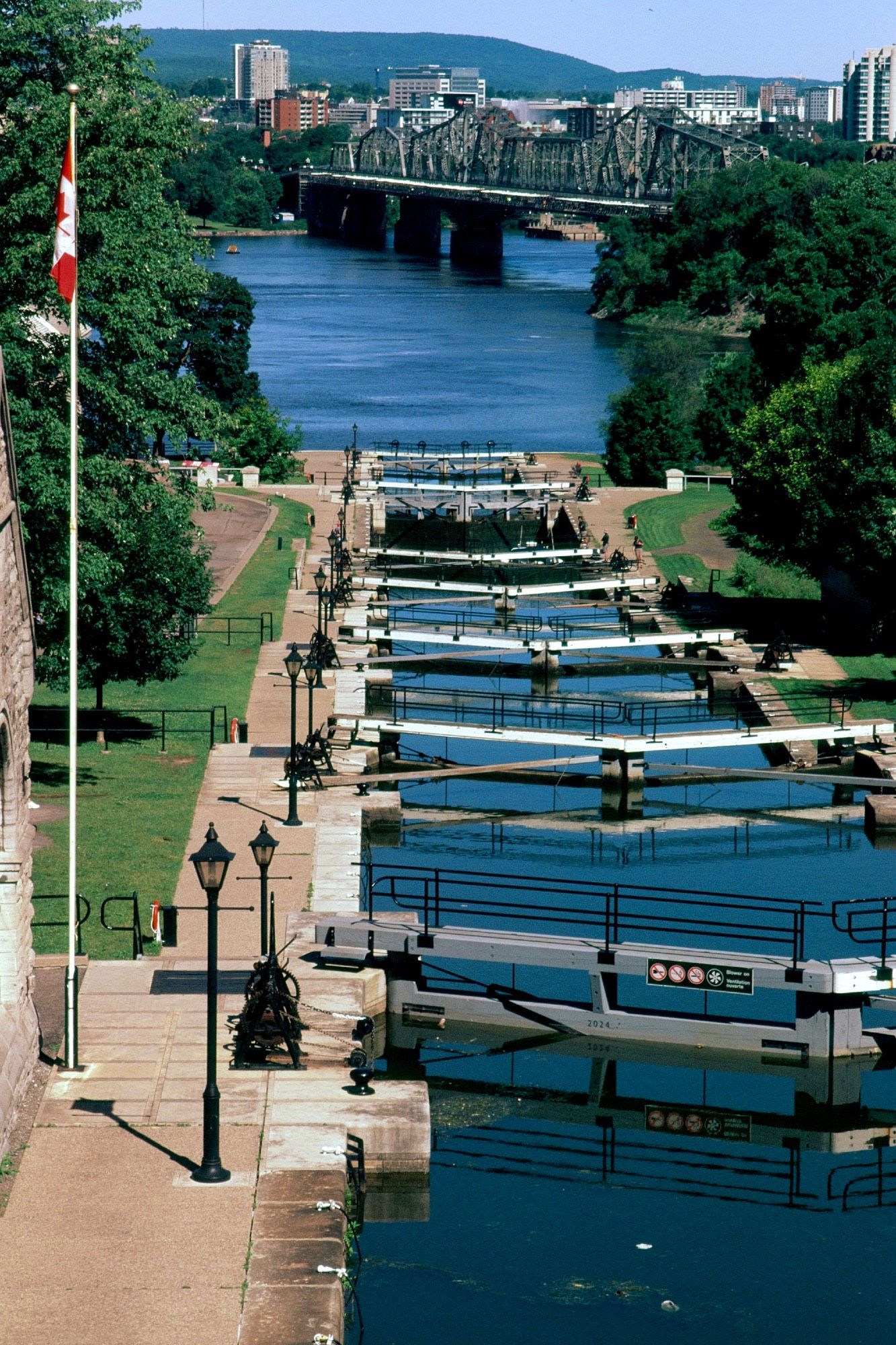 View of a set of canal locks from an elevated position. The canal leads down to a wide and calm river (the Ottawa River). In the background is a steel bridge to the city across the river, Hull, Quebec.