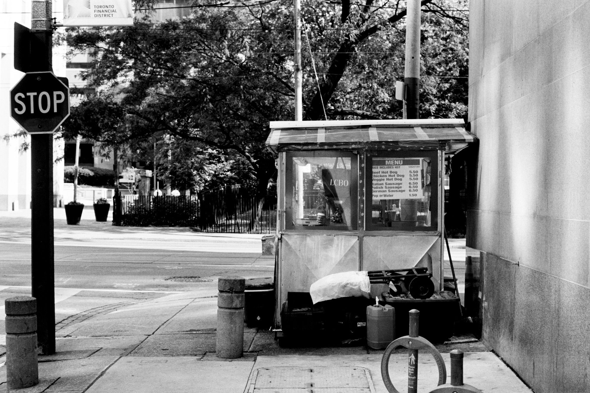 Hot dog cart parked beside a stone clad building at opening in the morning. Its attendant is inside reading her phone and enjoying her morning coffee.