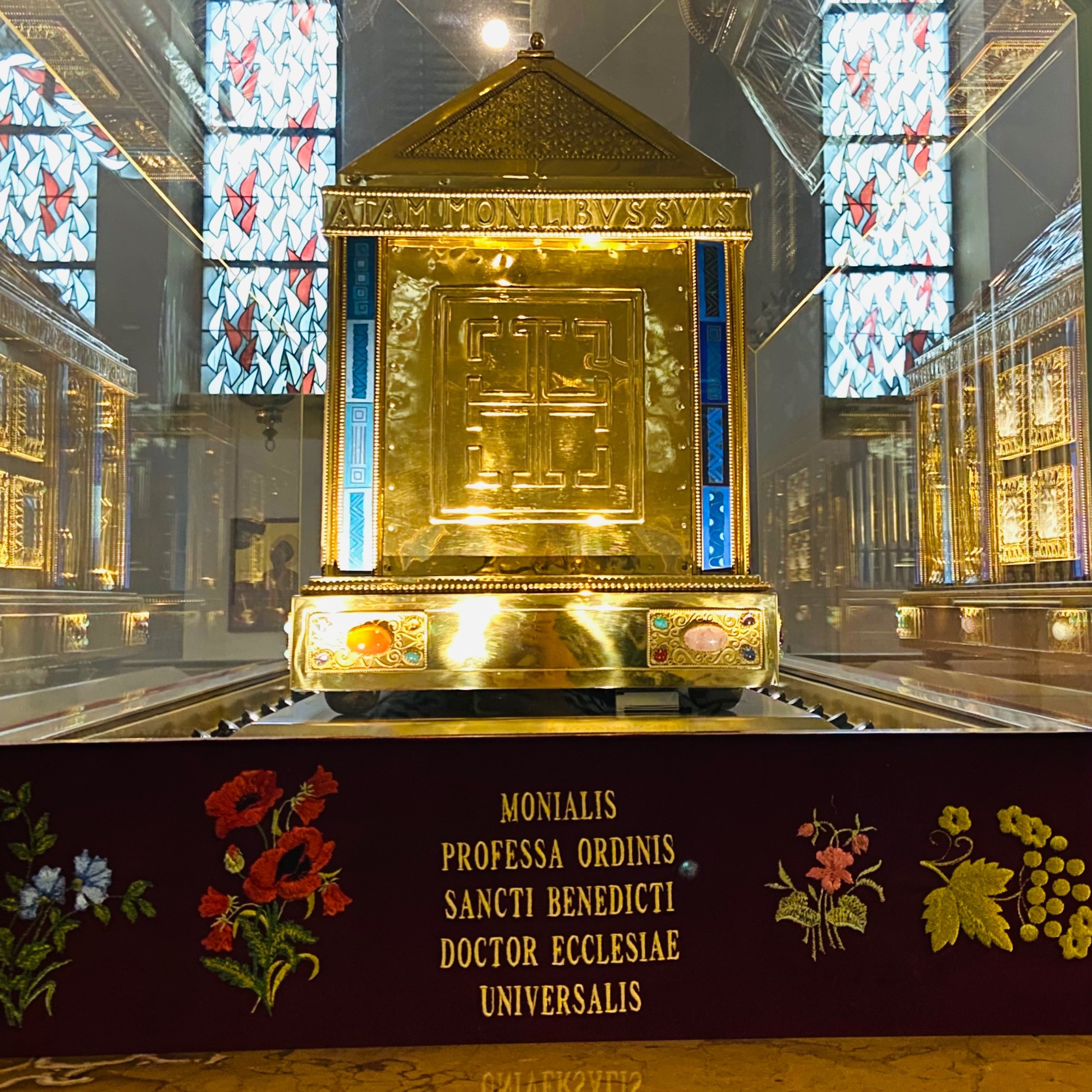 Side view of golden reliquary and below it in Latin and surrounded by embroidered flowers - “monialis professa ordinis sancti benedicti doctor ecclesiae universalis”