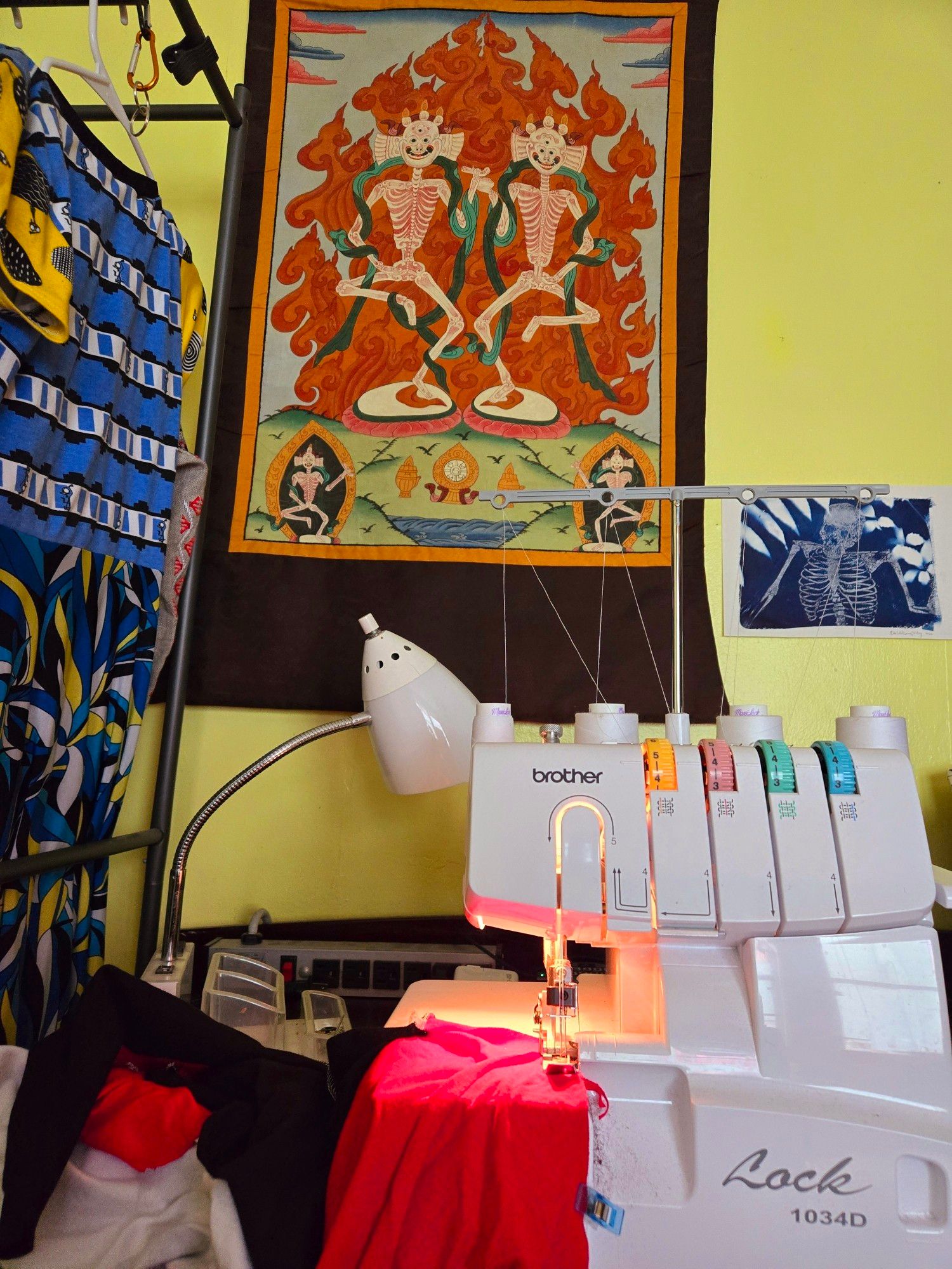 A serger sewing red fabric. In the background, a Tibetan scroll painting of dancing skeletons and a cyanotype print of a skeleton.