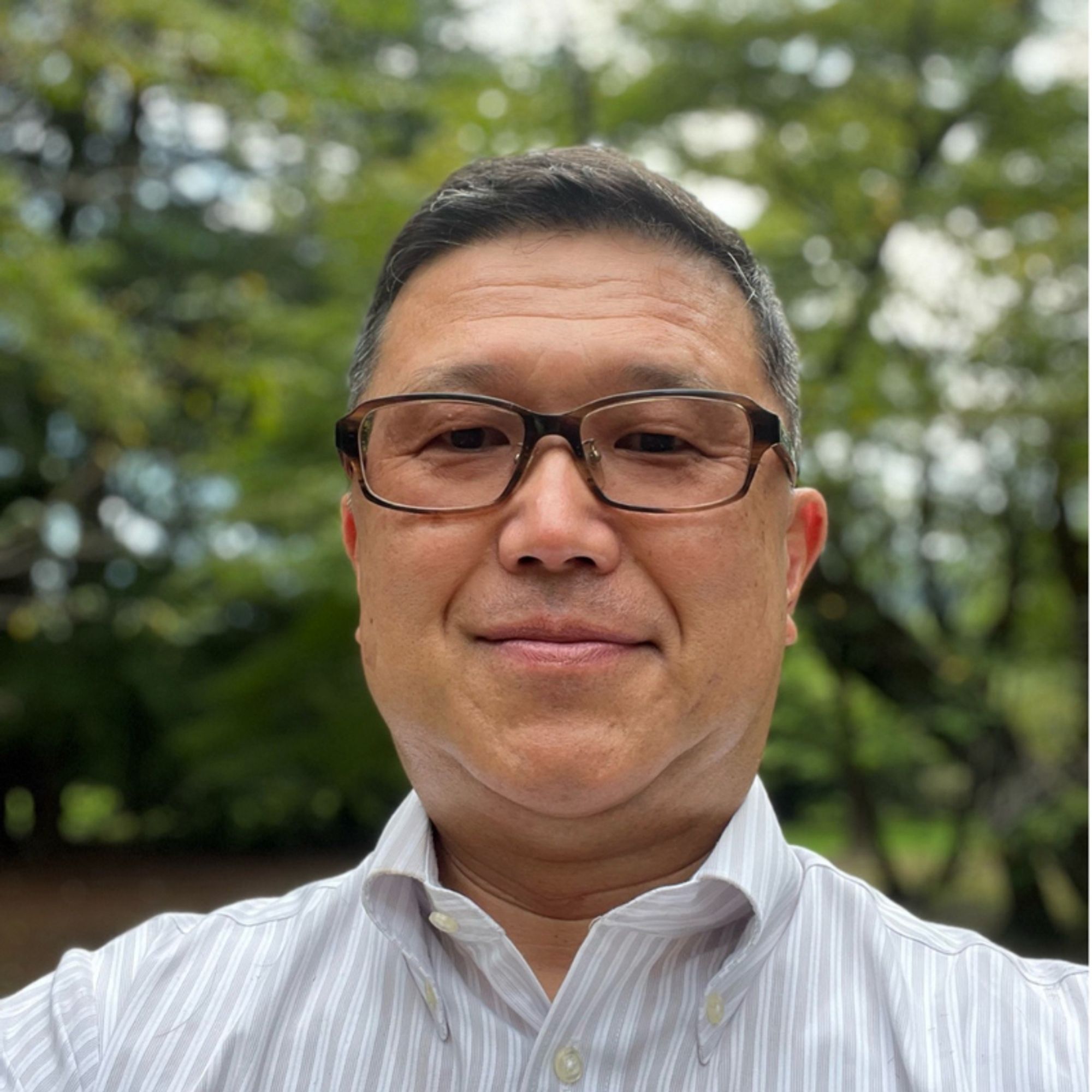 A man wearing glasses and a light-colored, striped button-down shirt smiles slightly at the camera. He has short, dark hair and is standing outdoors, with a background of blurred trees and greenery