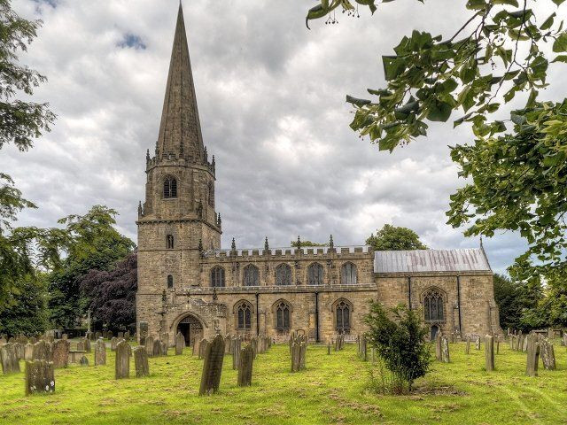 Photo of a church with a spire. Green graveyard with standing stones in foreground and cloudy sky above