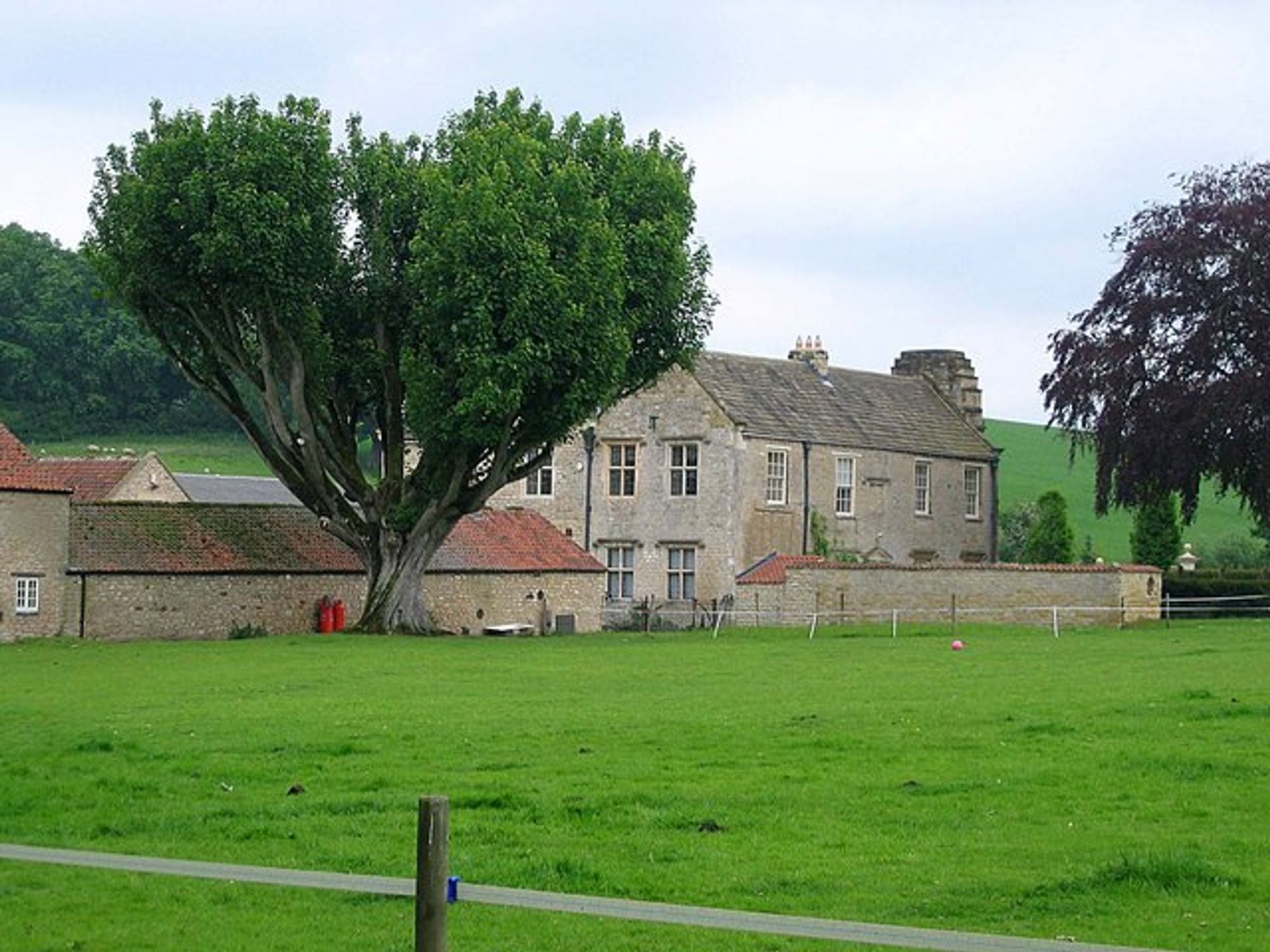 Photo of an old house with lower farm buildings to the left. Taken from across a very green field with a large tree in front of the buildings.