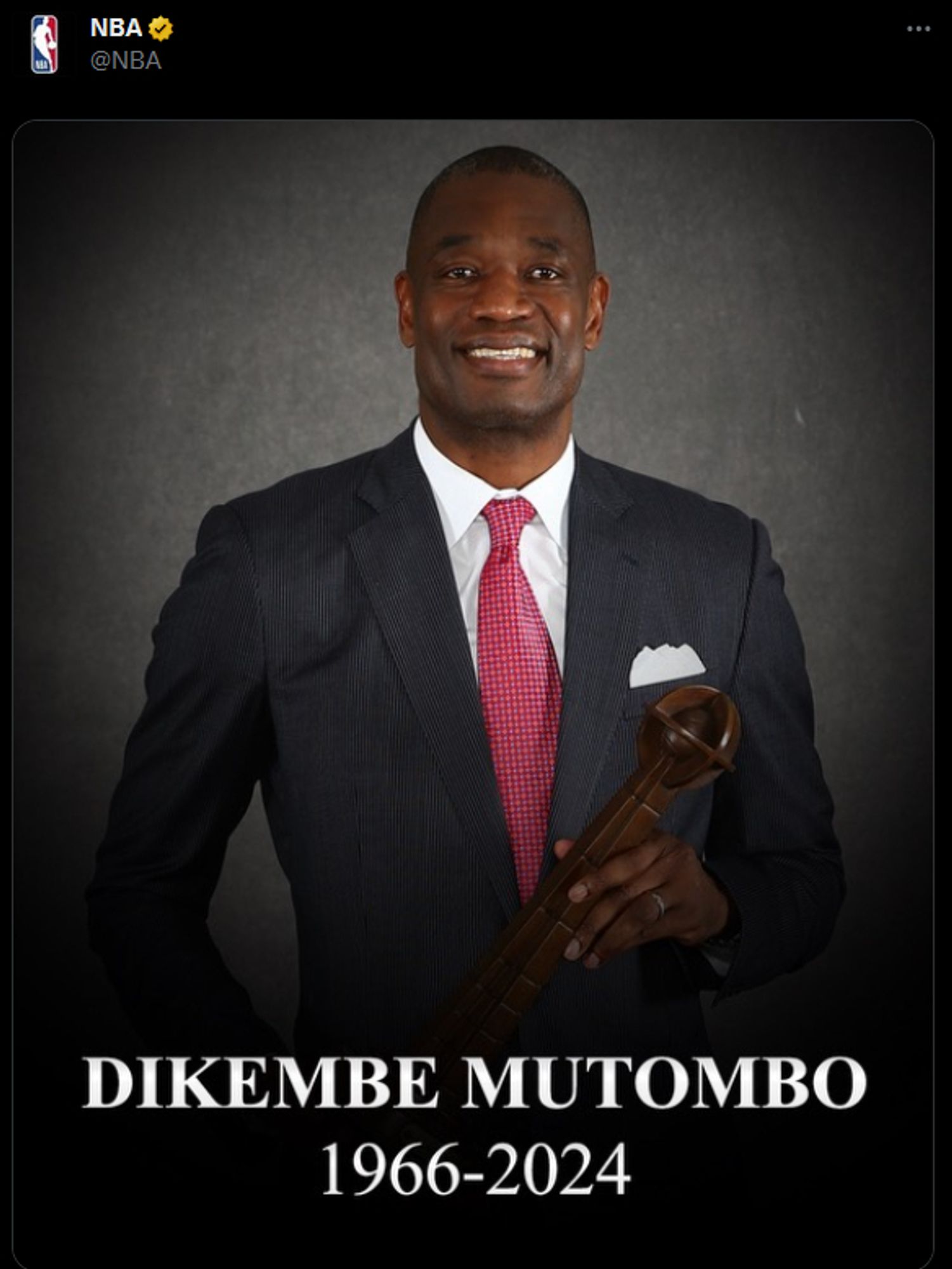 The tweet from the NBA. Same as above, but Mutombo's head is not cut off by the framing. He's smiling and holding a wooden staff. He's wearing a suit and red tie with white cross hatching.