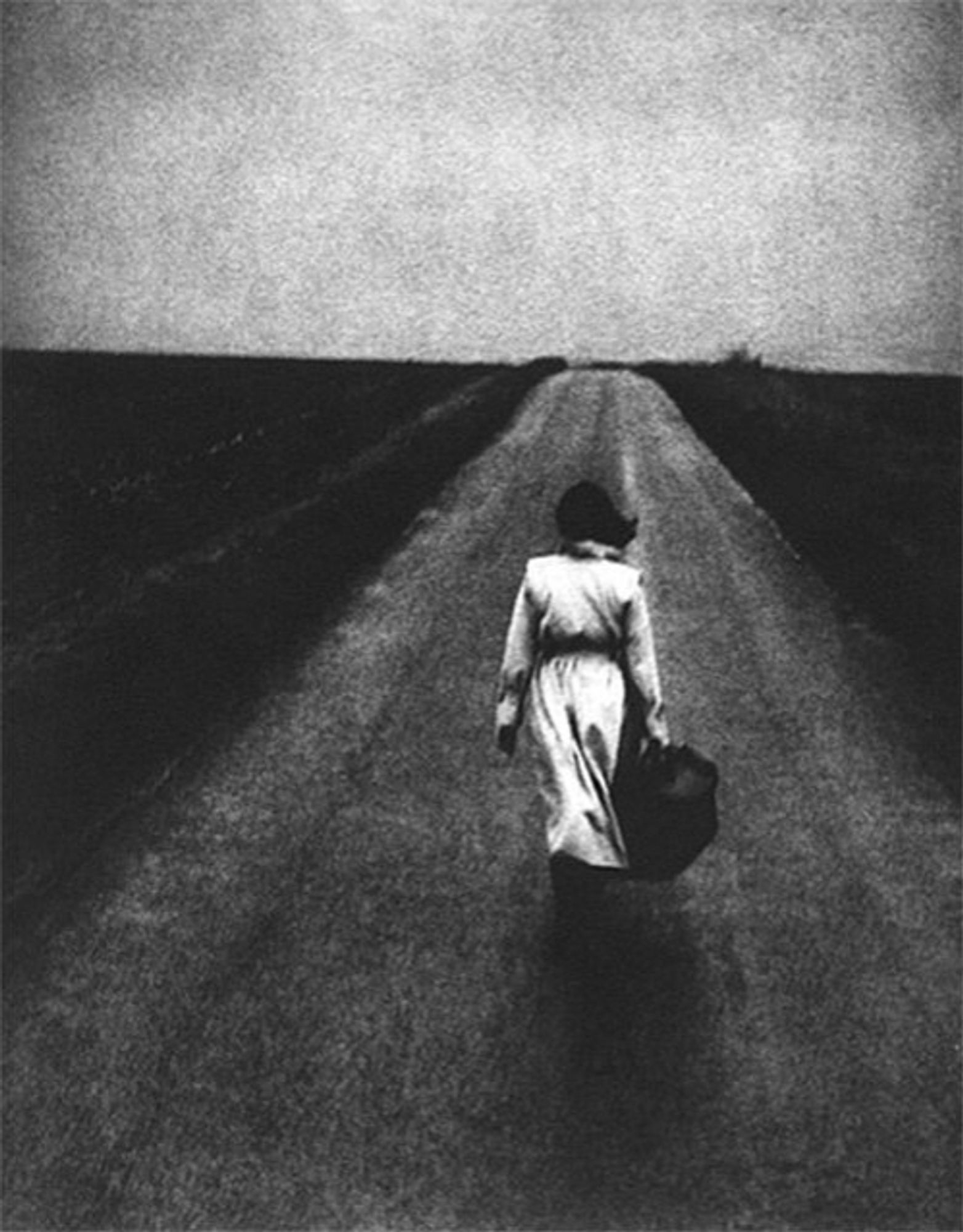 'Road. East of England.'  Woman in a belted trench coat and carrying a bag is walking toward the horizon.