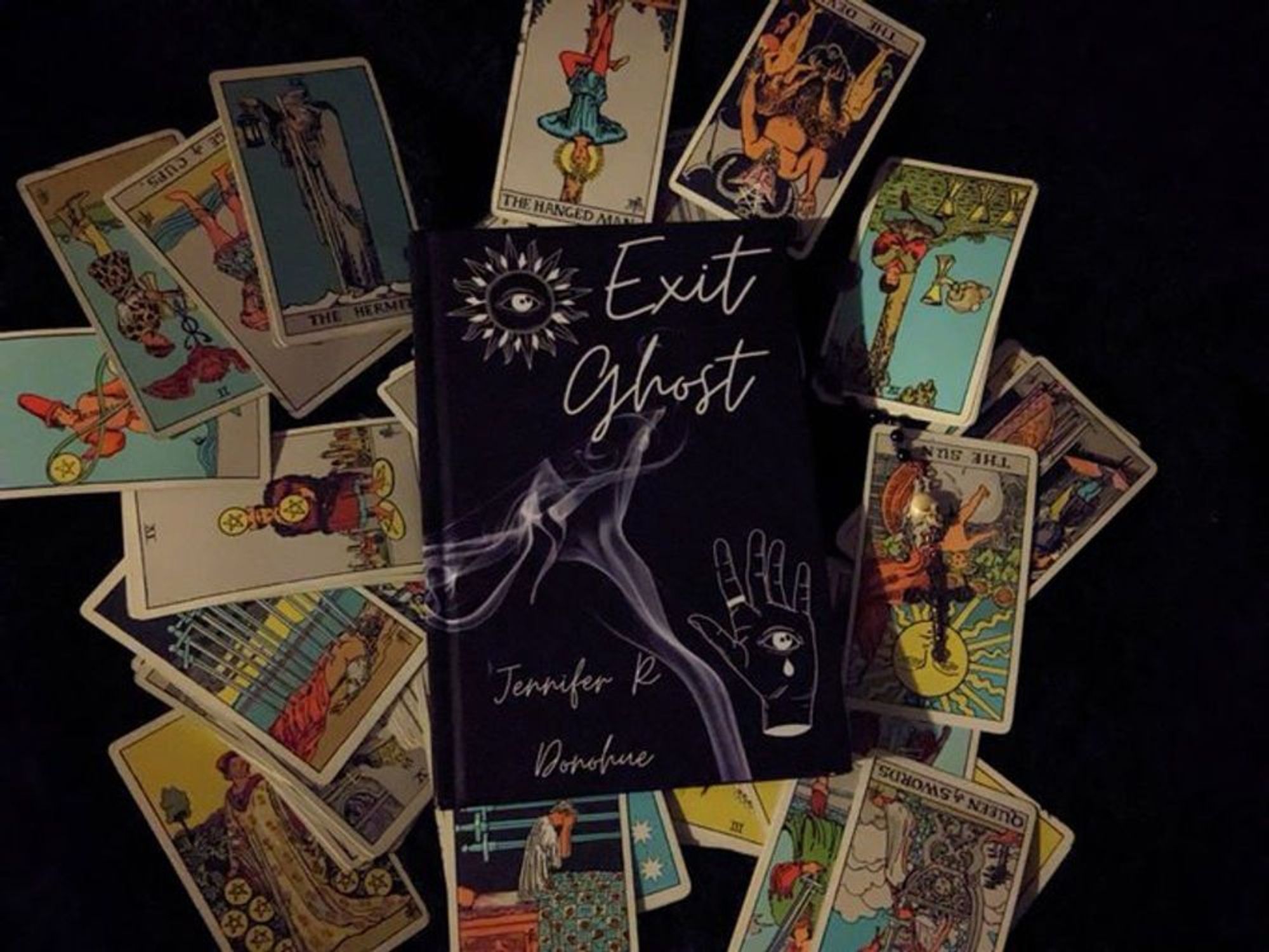 hardcover of Exit Ghost on a splayed out Rider Waite tarot deck, with a single-decade rosary that has a bone skull as a focal bead above the cross  

  (Exit Ghost book cover: a black background with a wisp of smoke curling up, a sketched/stylized sun with an eye in the middle of it in the upper left corner, a sketched hand in the lower right corner wearing a ring in the first finger, with the same eye in the palm, with a single tear falling from it, "Exit Ghost" above it in a cursive/handwrite-y font, Jennifer R. Donohue to the bottom left in the same font. )

I designed this cover in Canva.