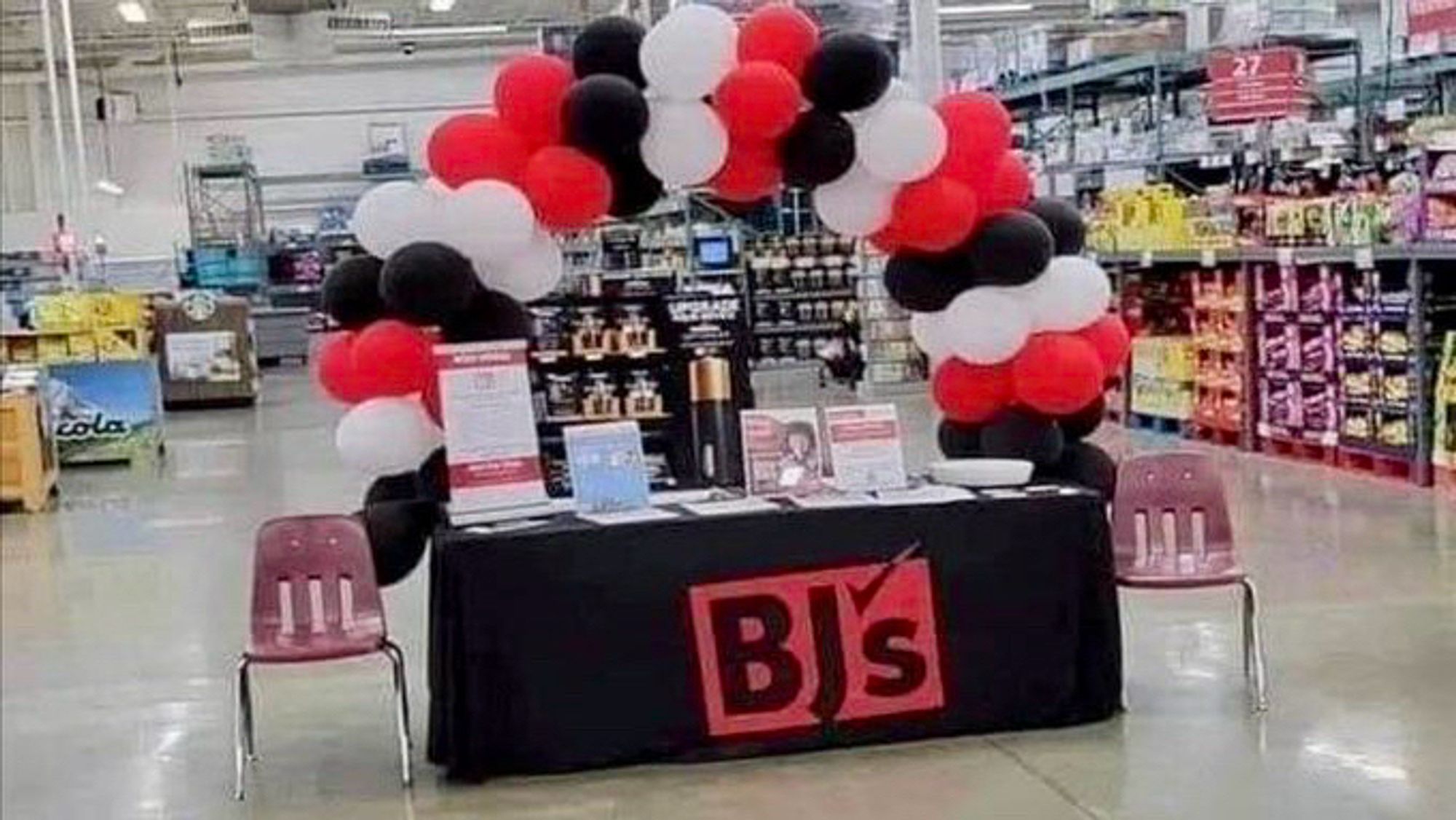 A promotional booth featuring a black tablecloth with the logo "BJ's" is set up in a store. Above the table is an arch made of red, black, and white balloons. Two empty chairs are placed in next to the table.