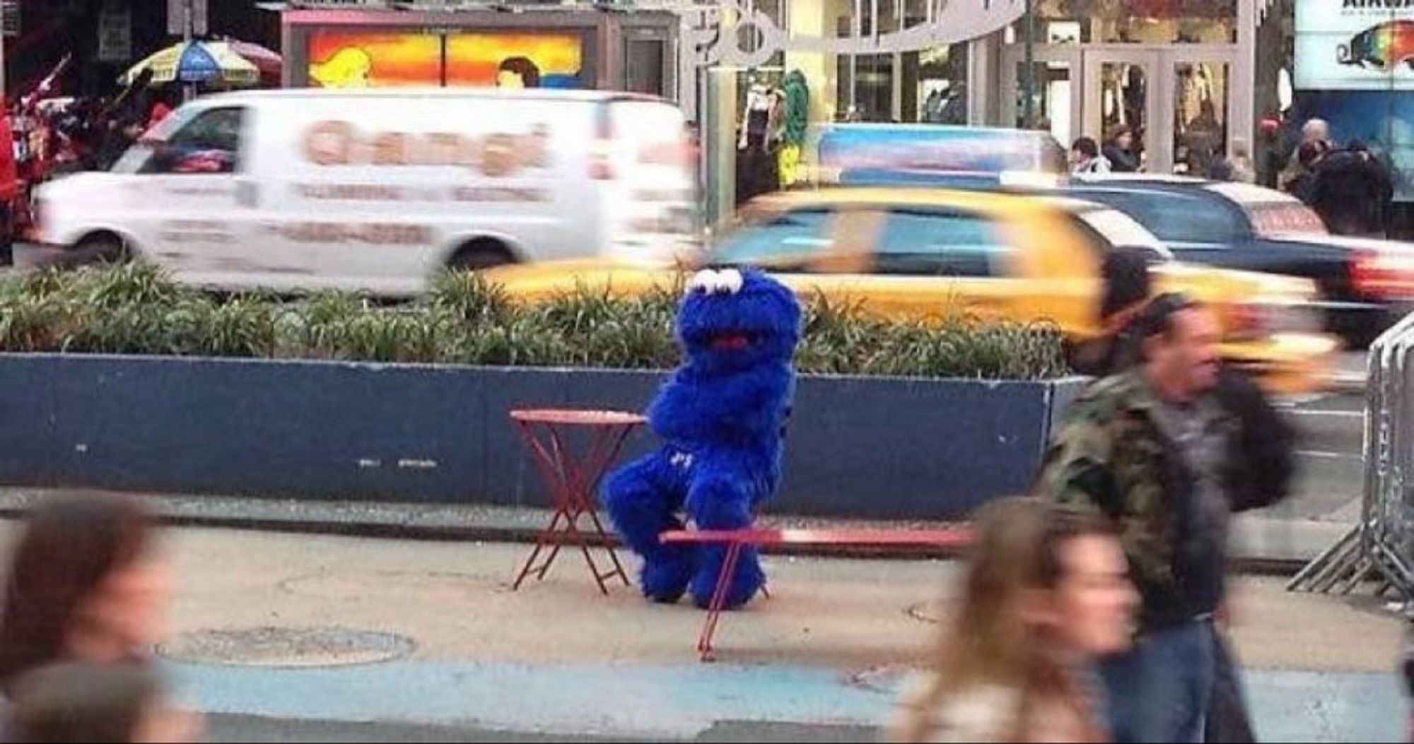 Cookie Monster sits at a sidewalk table as pedestrians and vehicles go by. His arms are crossed and he looks contemplative