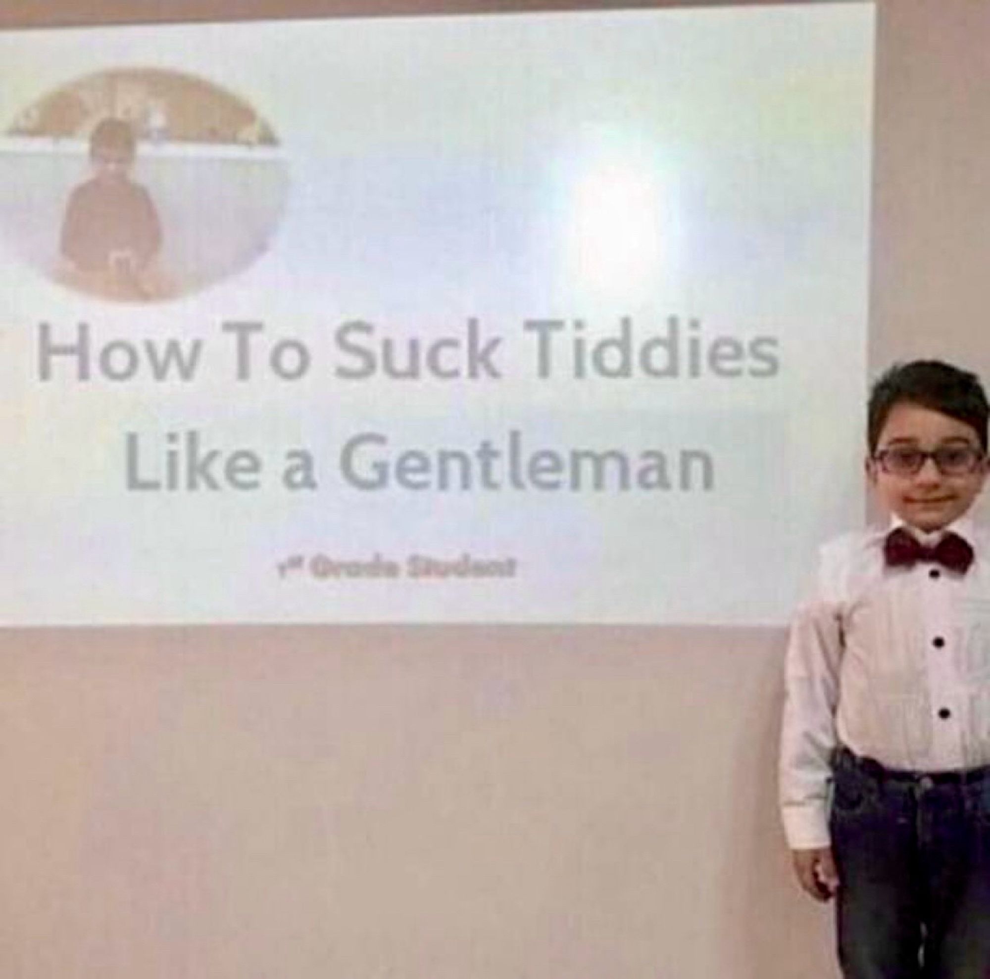 A smiling young boy in glasses and a bow tie stands next to a projection that reads, "How To Suck Tiddies Like a Gentleman, 1st Grade Student."