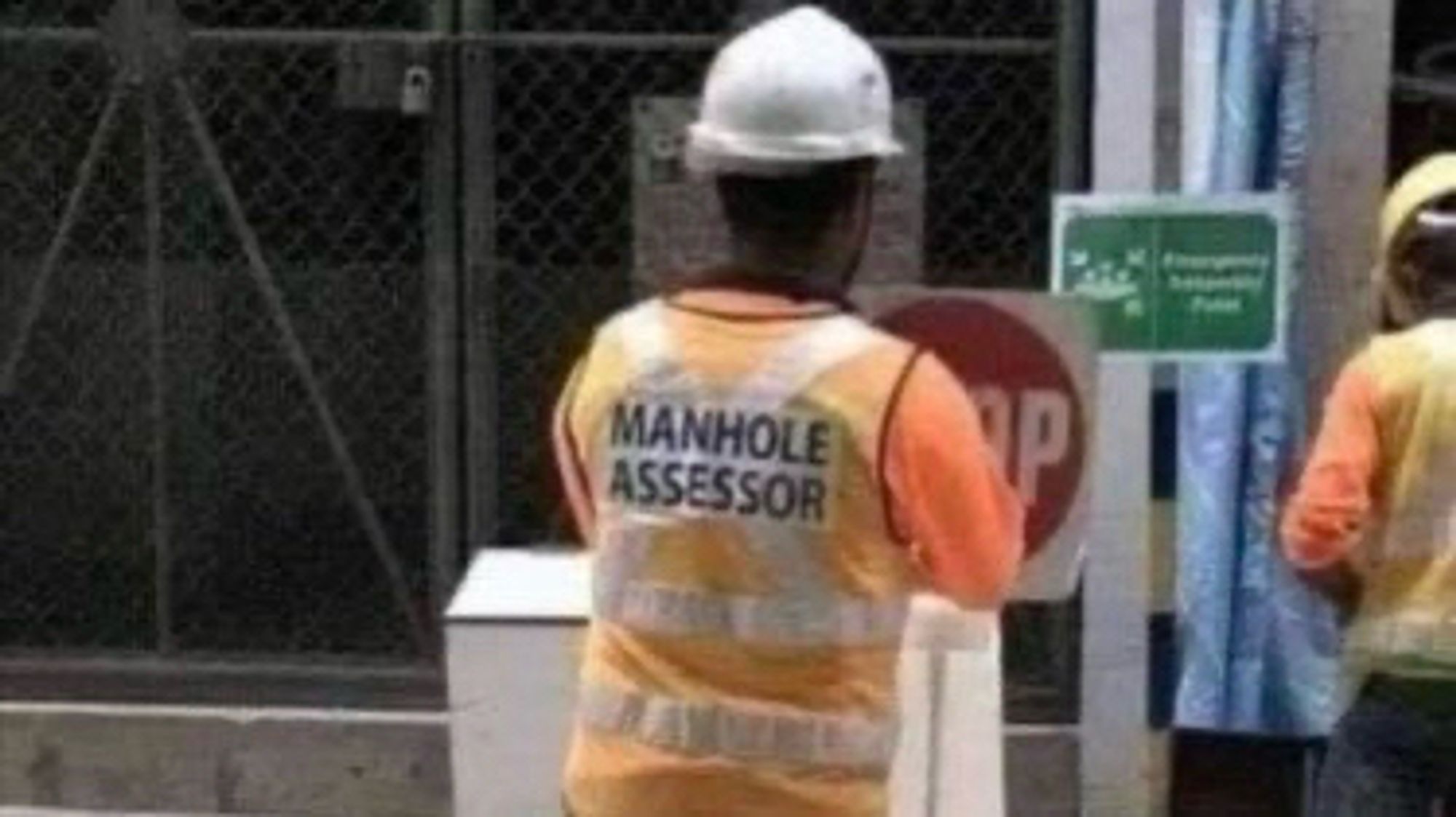 A construction worker wearing a safety helmet and a high-visibility vest labeled "MANHOLE ASSESSOR" stands in front of a stop sign and a green informational sign.