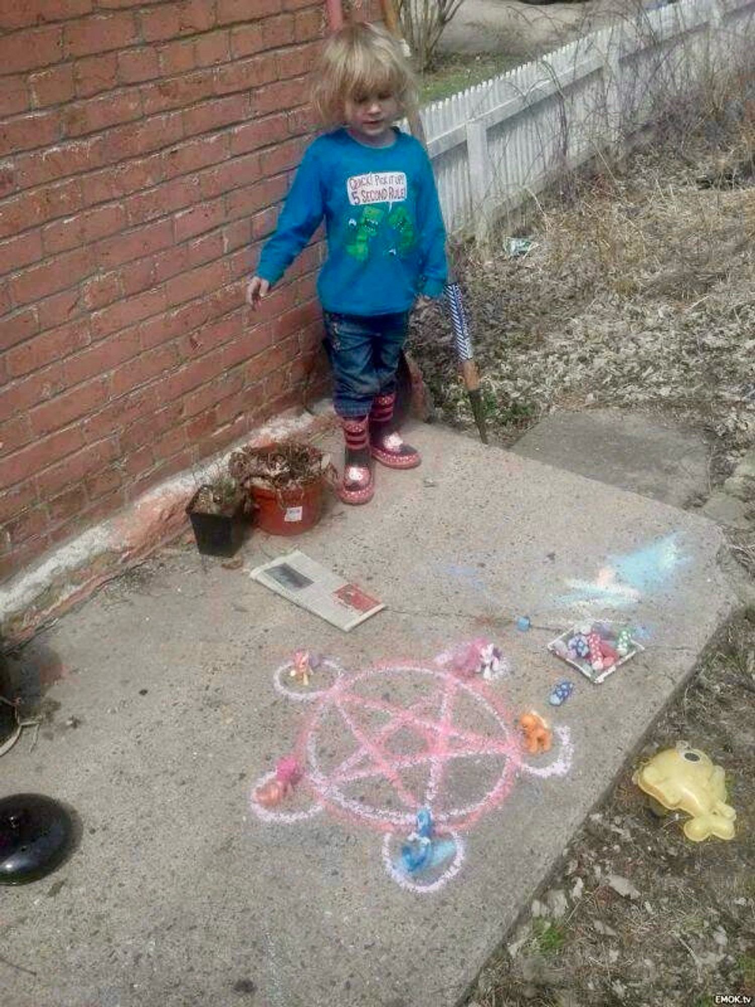 A child stands above a colorful chalk drawing of a pentagram on a concrete surface, small My Little Pony toy figures sit at each point.