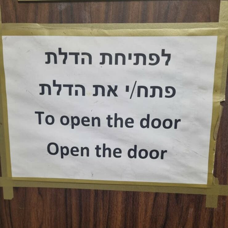 Sign taped to a door has two lines in Hebrew, followed underneath by "To open the door - Open the door" in English.