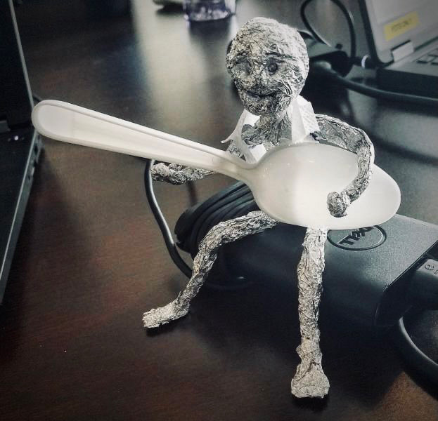 A small sitting figure made from tin foil holds a spoon like a guitar. 