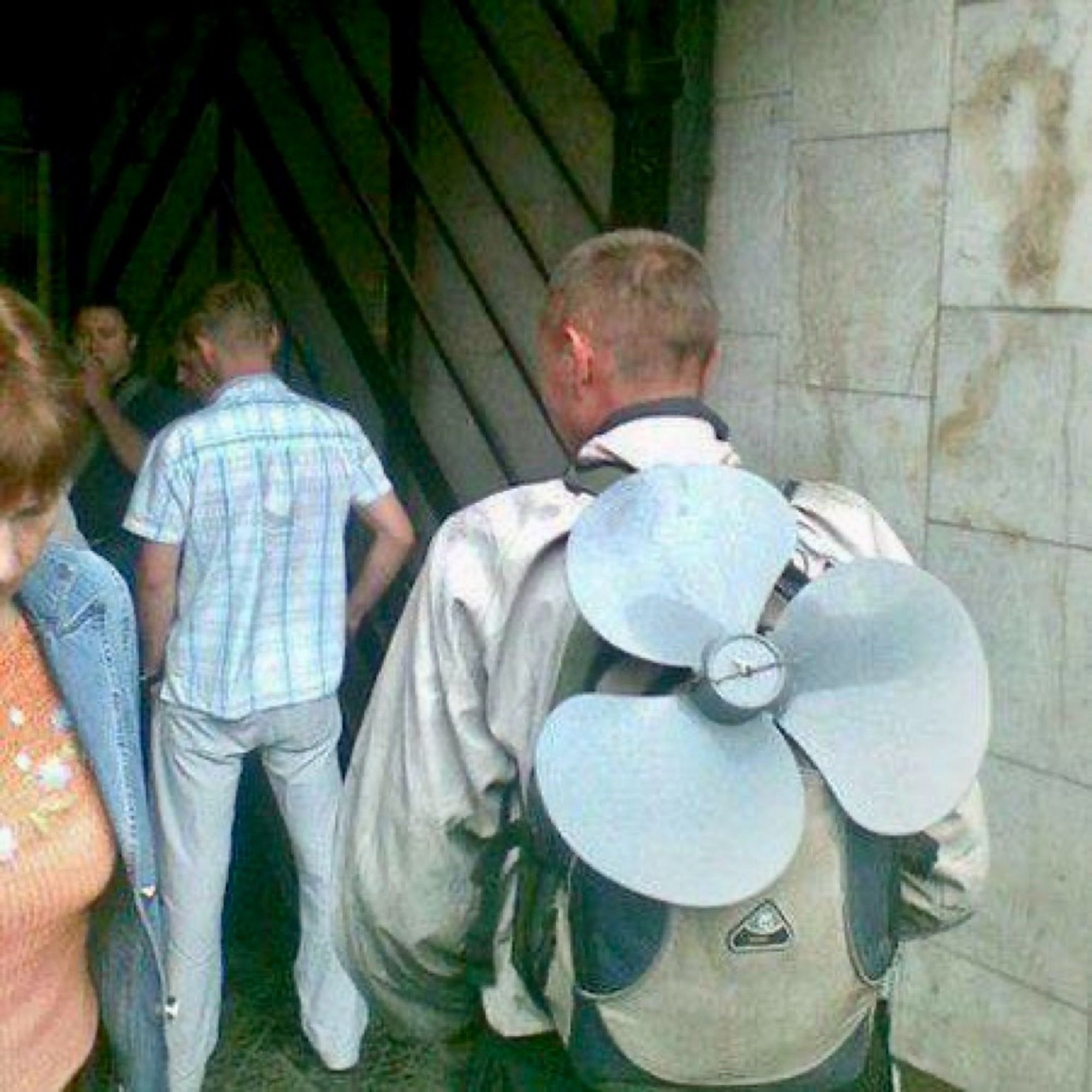 A person stands in a small crowd wearing a backpack with three large fan blades protruding from it.